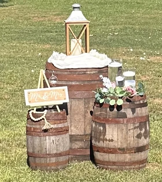 Decorative outdoor setup with wooden barrels, glass jars, a lantern, and a floral arrangement for a wedding or event.
