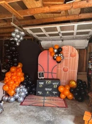 Indoor basketball-themed photo setup with orange, black, and silver balloons, a mini basketball hoop and backboard, a chalkboard sign, and a red and white rug.