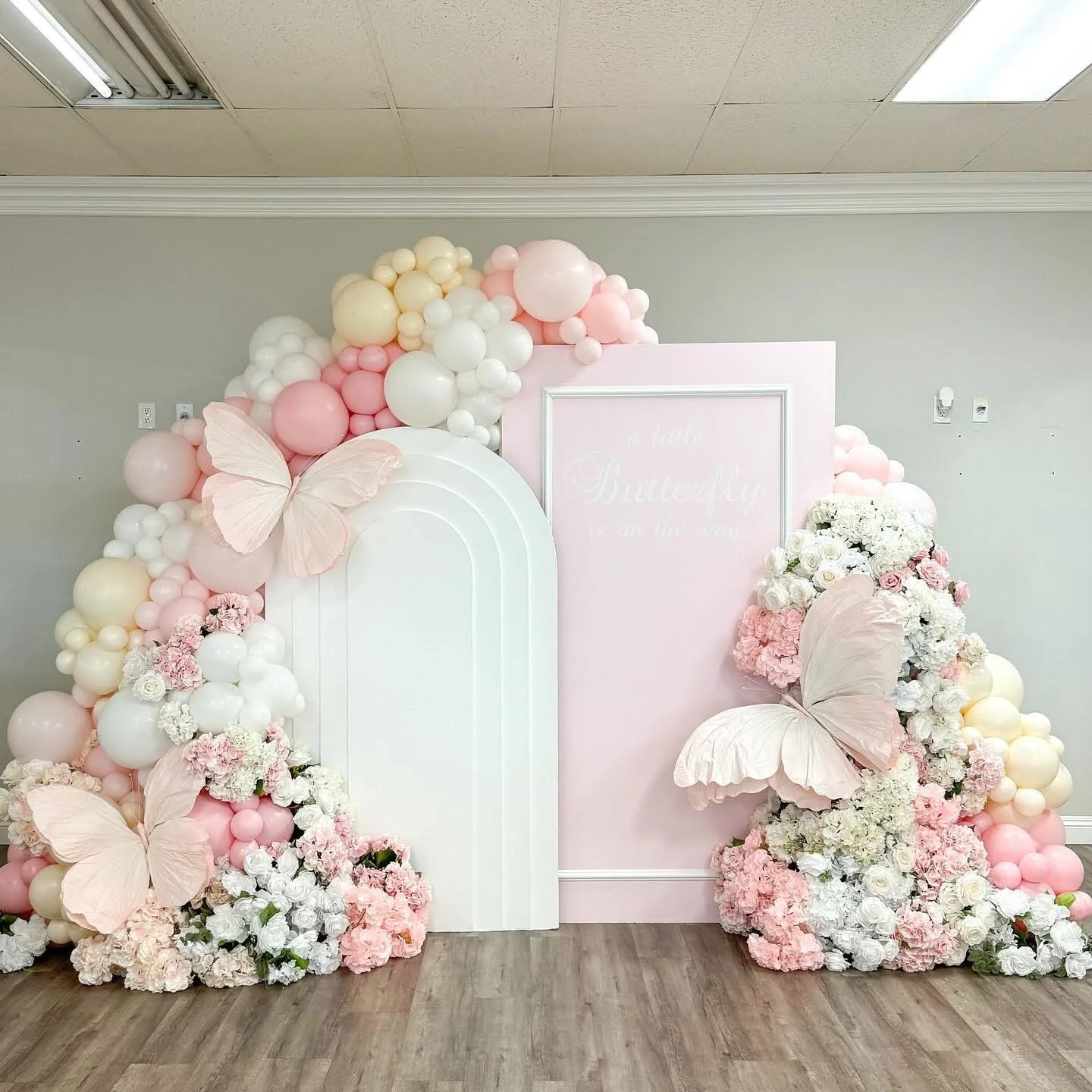Decorative flower and balloon arrangement with pink, white, and cream-colored flowers, large paper butterflies, and a pink and white backdrop with a partially visible sign.