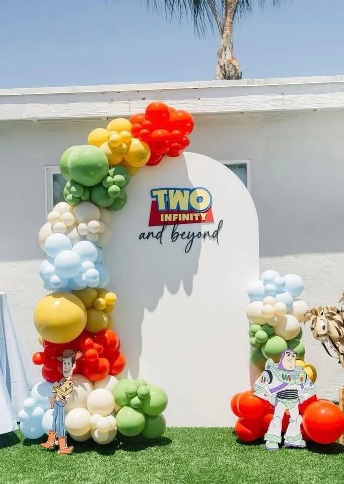 Decorative display with balloons in rainbow colors surrounding a white backdrop featuring the 'Toy Story' logo and characters Woody and Buzz Lightyear, with a palm tree and a blue sky in the background.