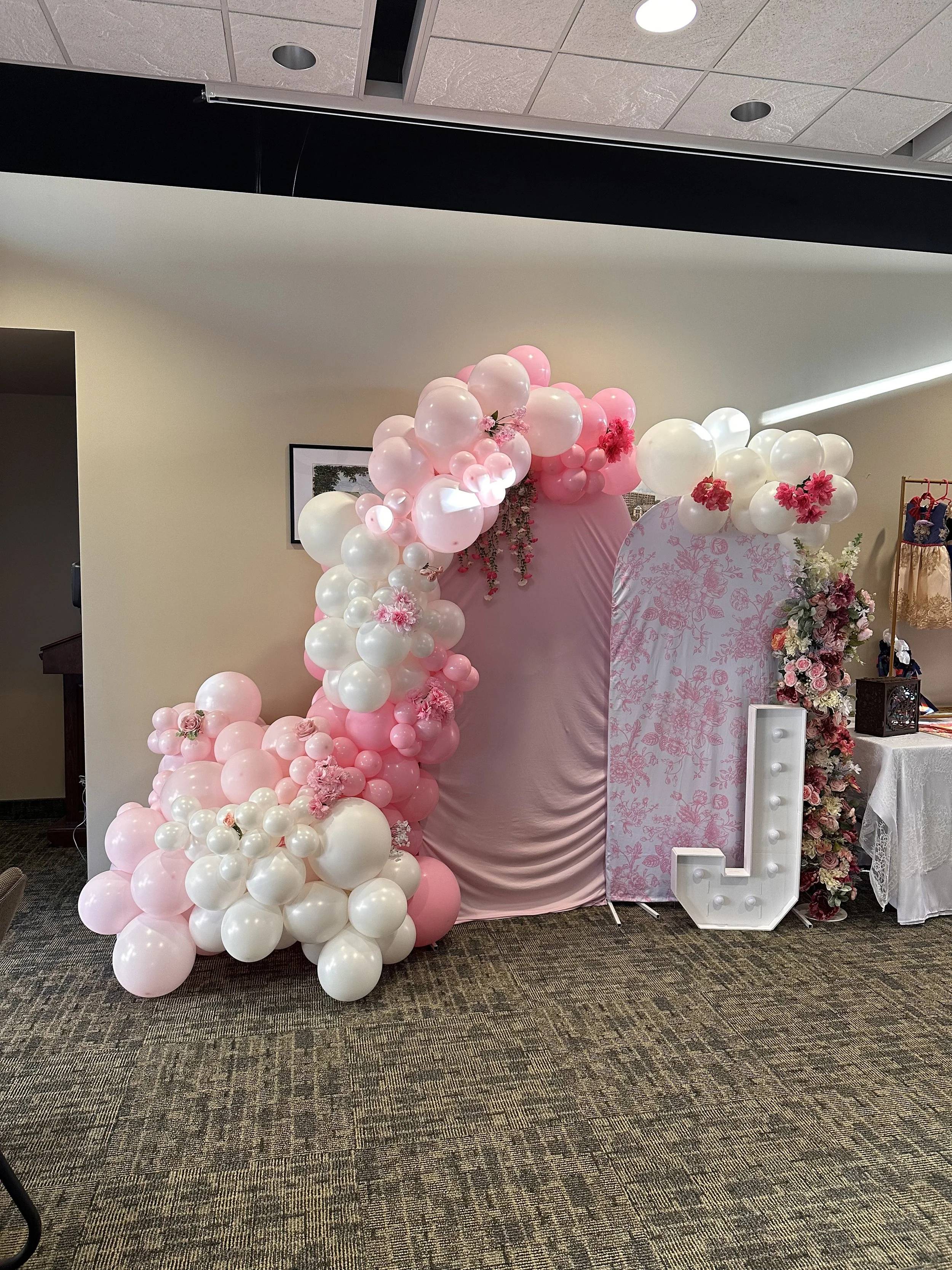 Pink and white balloon arch with floral decorations, a pink draped backdrop, and a large illuminated letter "L" at a decorated event setting.