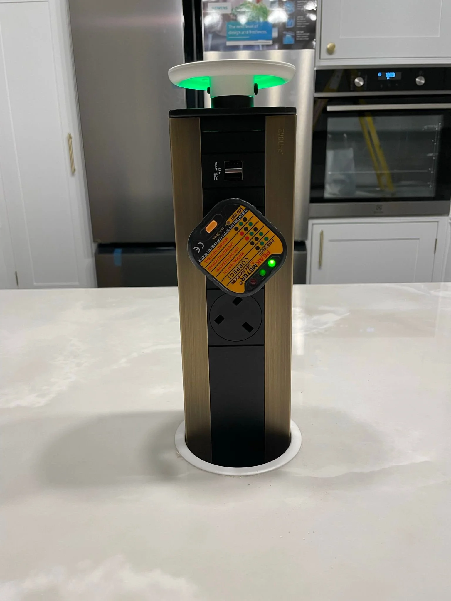 Kitchen countertop with a black and bronze power strip connected to a white smart device with green lighting, multiple outlets, and a control card hanging on it.