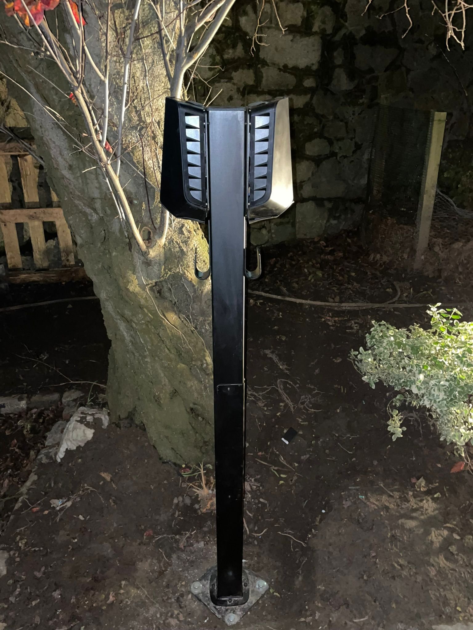 A tall black outdoor electric vehicle dual station in a garden area with a tree, rocks, and bushes around it at night.