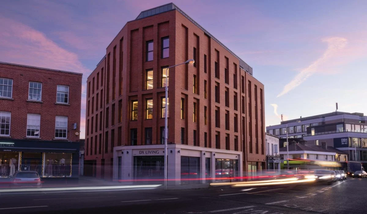 A multi-story modern brick building on a city street during dusk with light trails from moving vehicles in front.