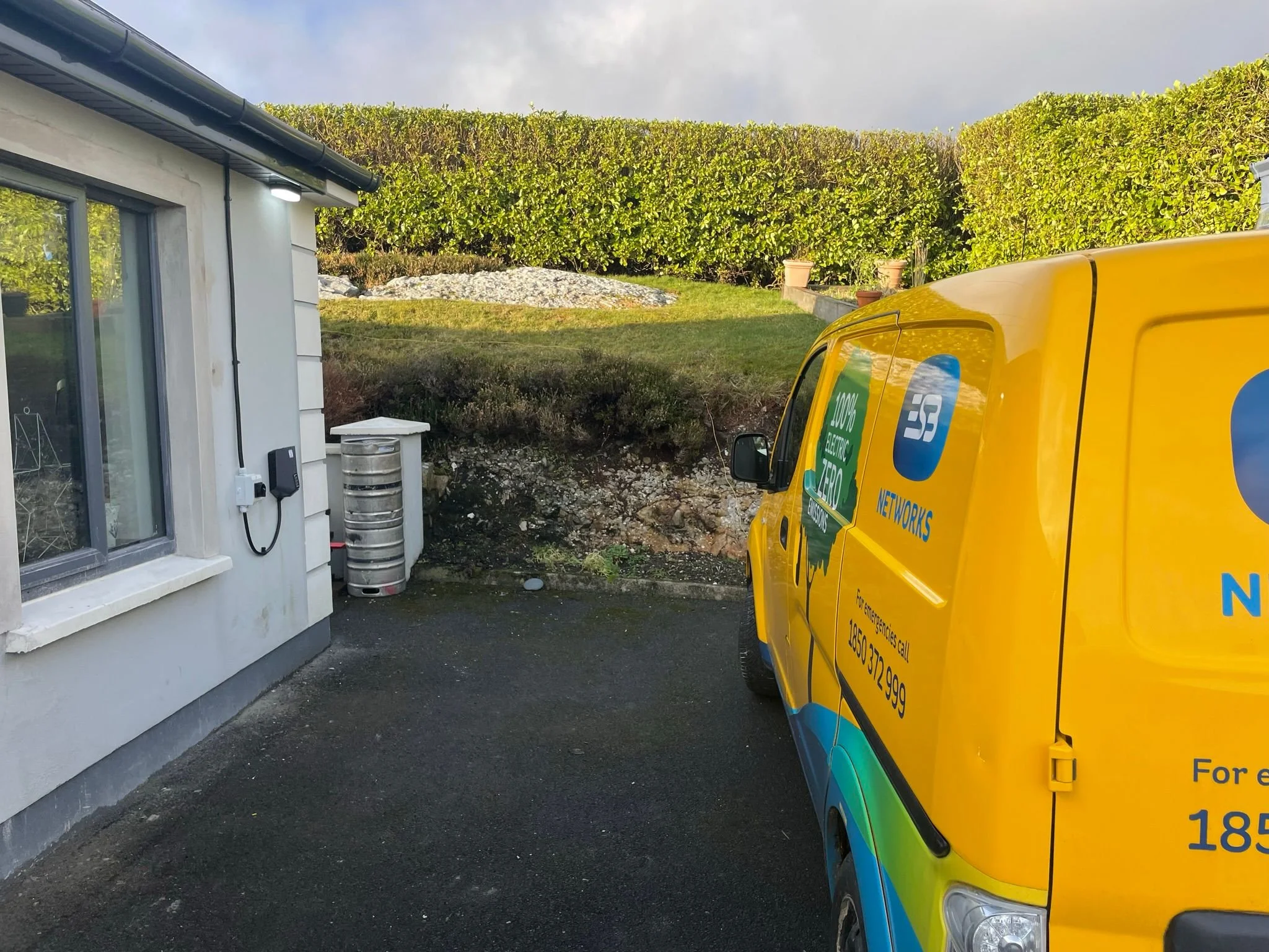 Yellow service van parked outside a white building with a window. A window air conditioning unit and electric meter are visible on the building. A grassy hill with bushes and potted plants is in the background.