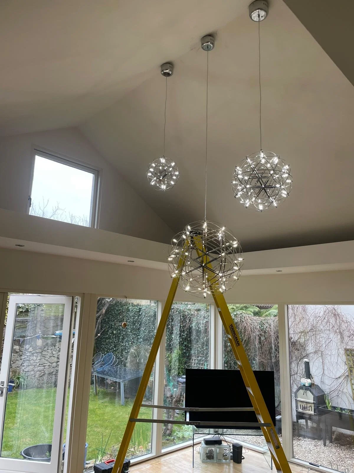 Interior view of a living room with modern chandelier fixtures hanging from the ceiling, a yellow ladder in the foreground, and large windows showing a backyard with grass, outdoor furniture, and trees.