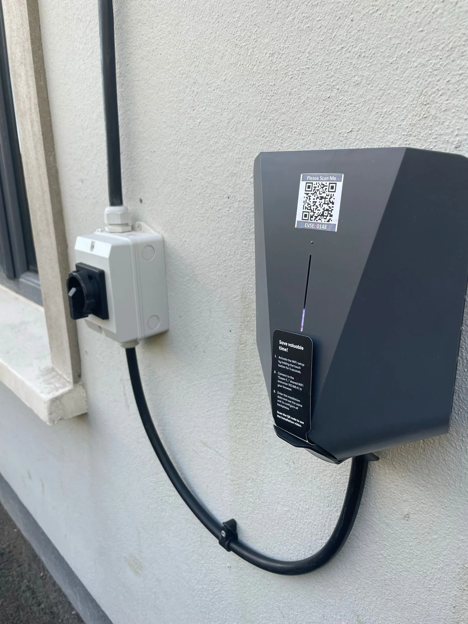 Electric vehicle charging station installed on a beige exterior wall, with a QR code label on top and a cord connected to a power outlet nearby.