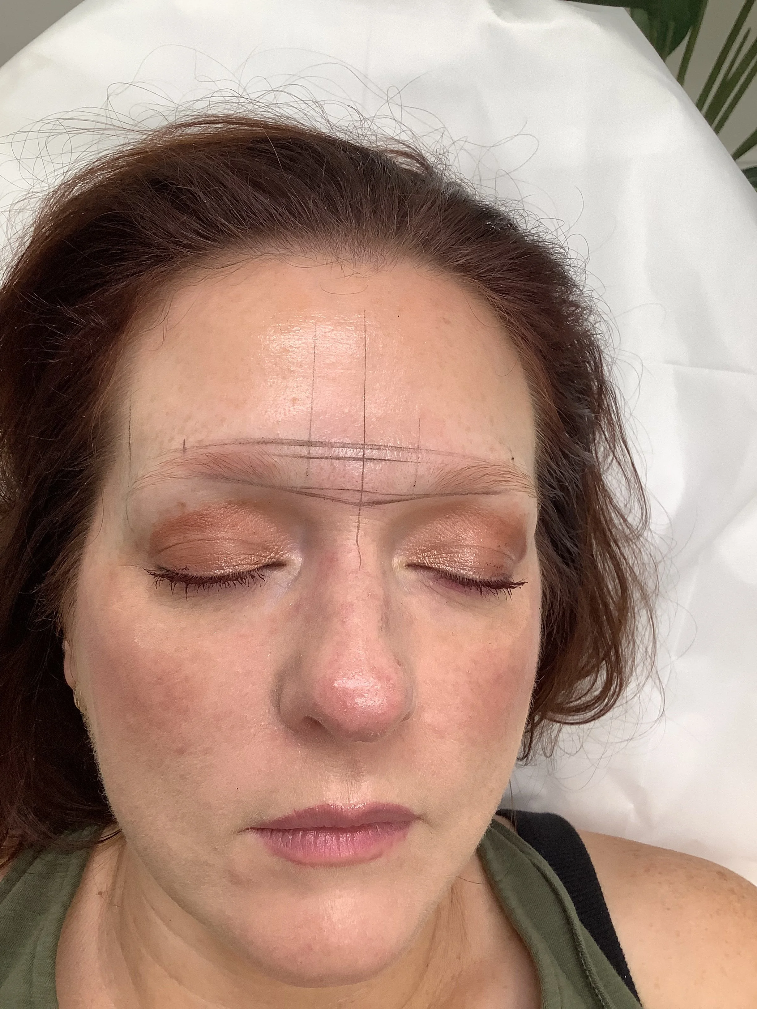Close-up of a woman with reddish-brown hair, closed eyes, and light makeup, lying on a white surface with markings on her forehead.