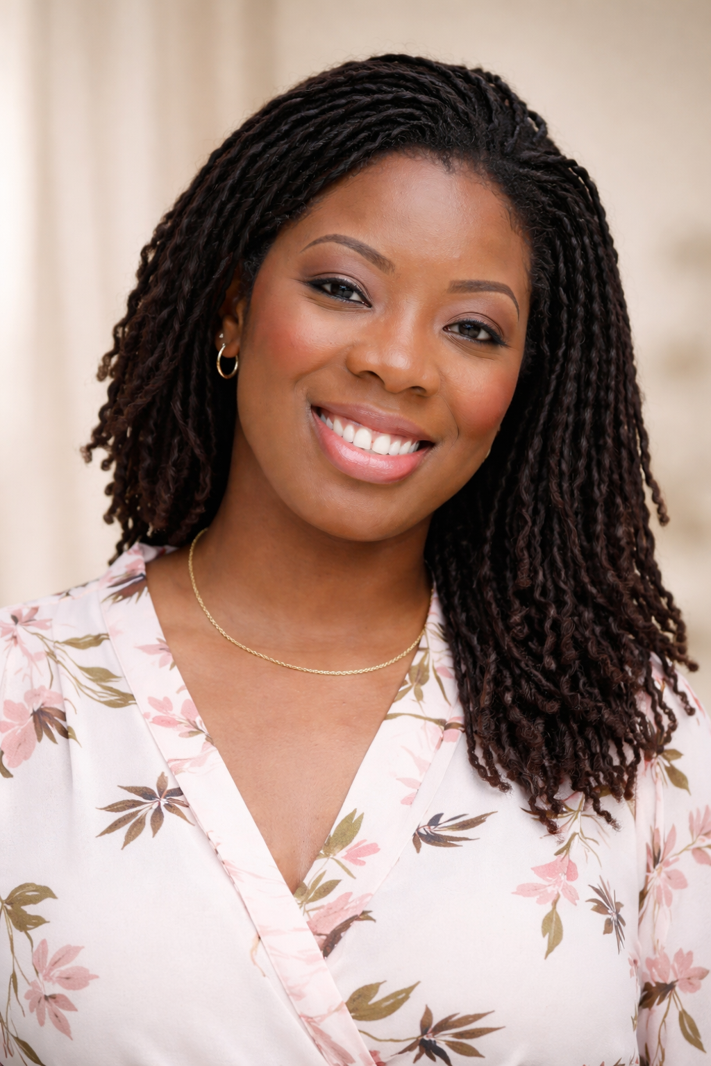 A smiling woman with dark, twisted hair and hoop earrings, wearing a floral blouse and a gold necklace.