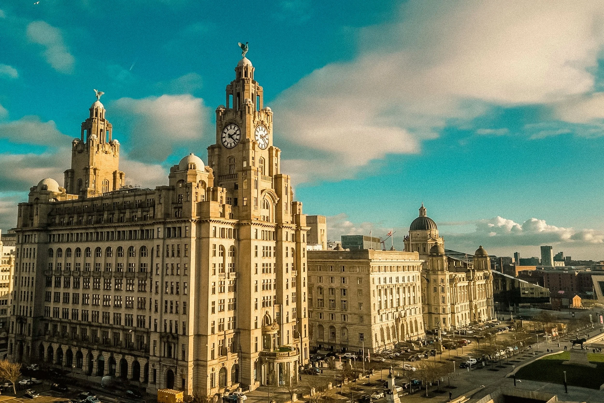 Two large historic buildings with clock towers, topped with statues of birds, under a partly cloudy sky in an urban setting.