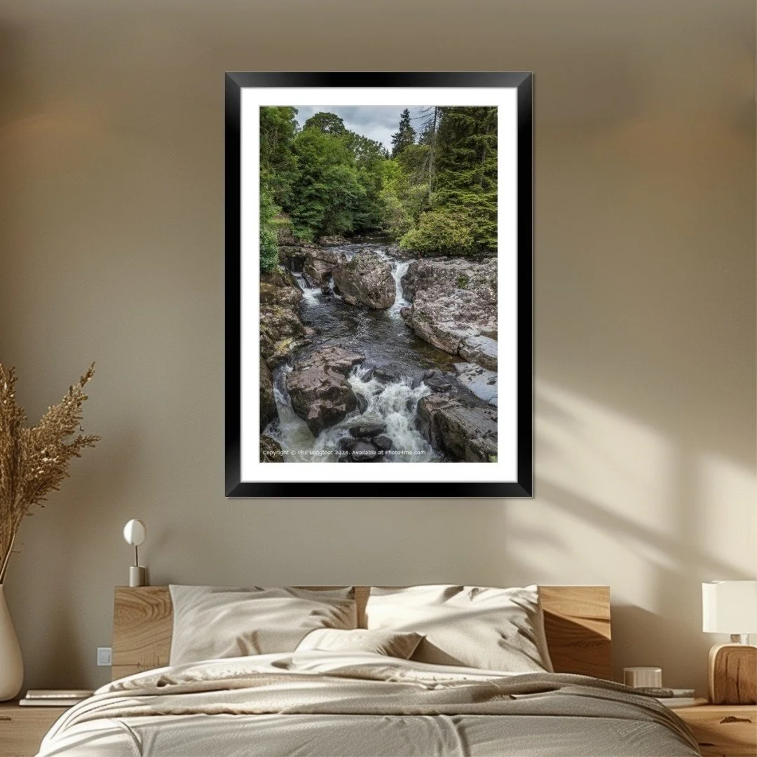 Framed landscape photograph of a rocky creek surrounded by lush green trees hanging on a beige bedroom wall above a bed with beige bedding