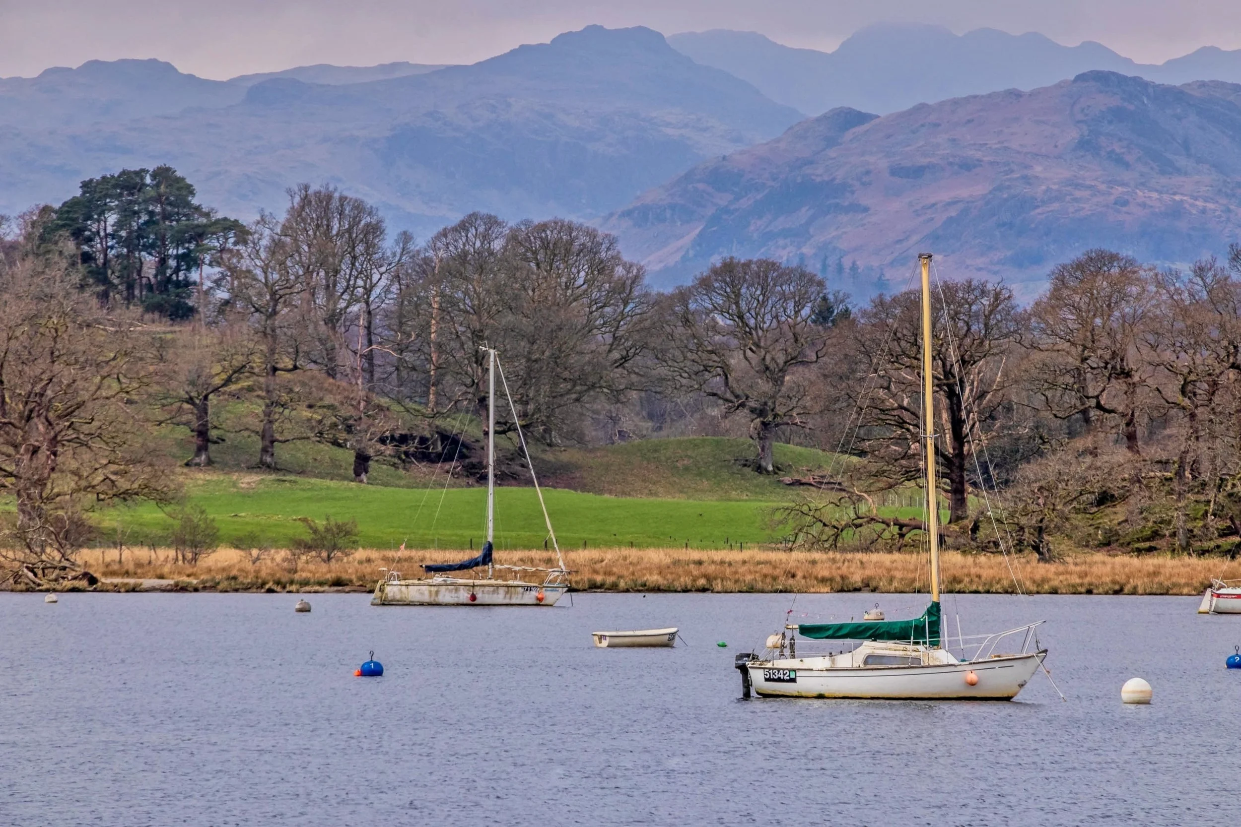 Sailing boats on a calm lake with a green shoreline, leafless trees, and mountains in the background on a cloudy day.
