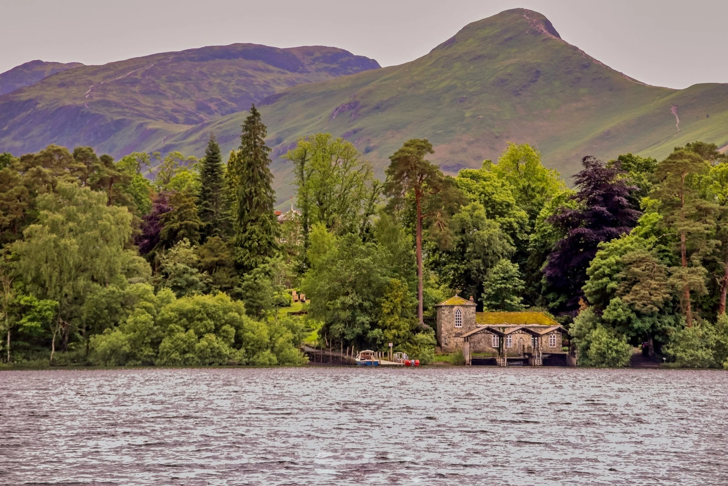 A body of water with small ripples in front of a lush green forested shoreline. Behind the trees, there are small buildings with stone walls and moss-covered roofs. Purple and green mountains rise in the background under an overcast sky.