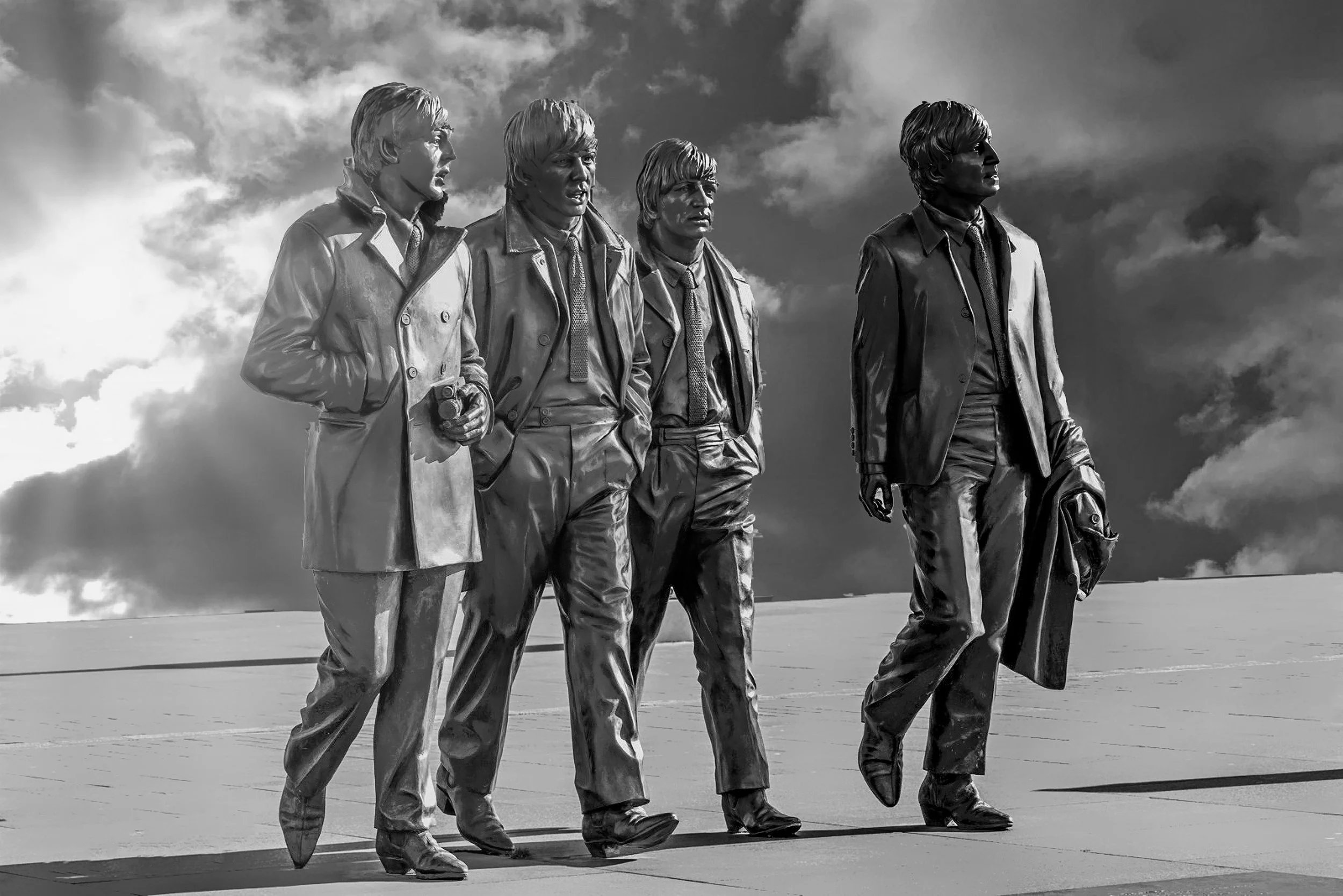 Black and white statue of four men walking, one holding a camera, with cloudy sky in the background.