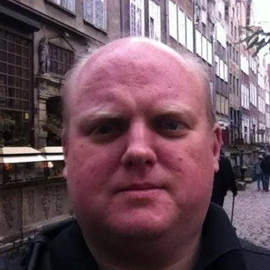 A close-up of a man with a shaved head standing outdoors on a city street with buildings and pedestrians in the background.
