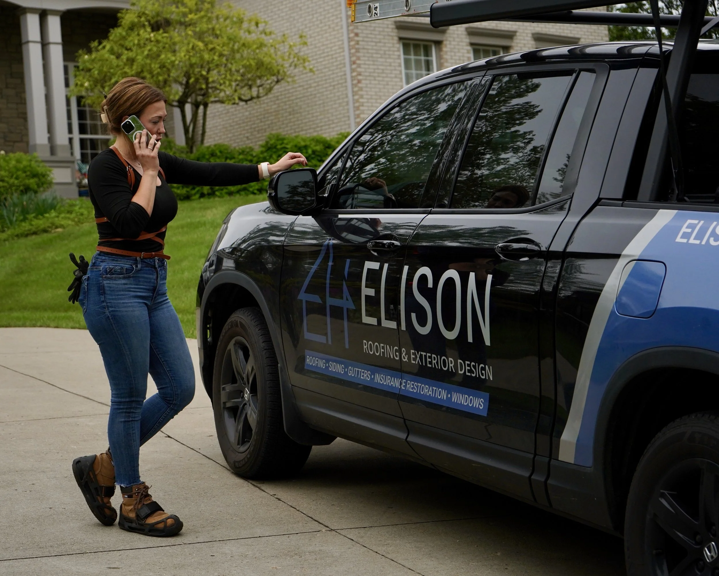 A woman talking on a cell phone while touching a car door handle of a black company vehicle with a logo for ELISON roofing and exterior design, parked on a driveway with a house and green lawn in the background.