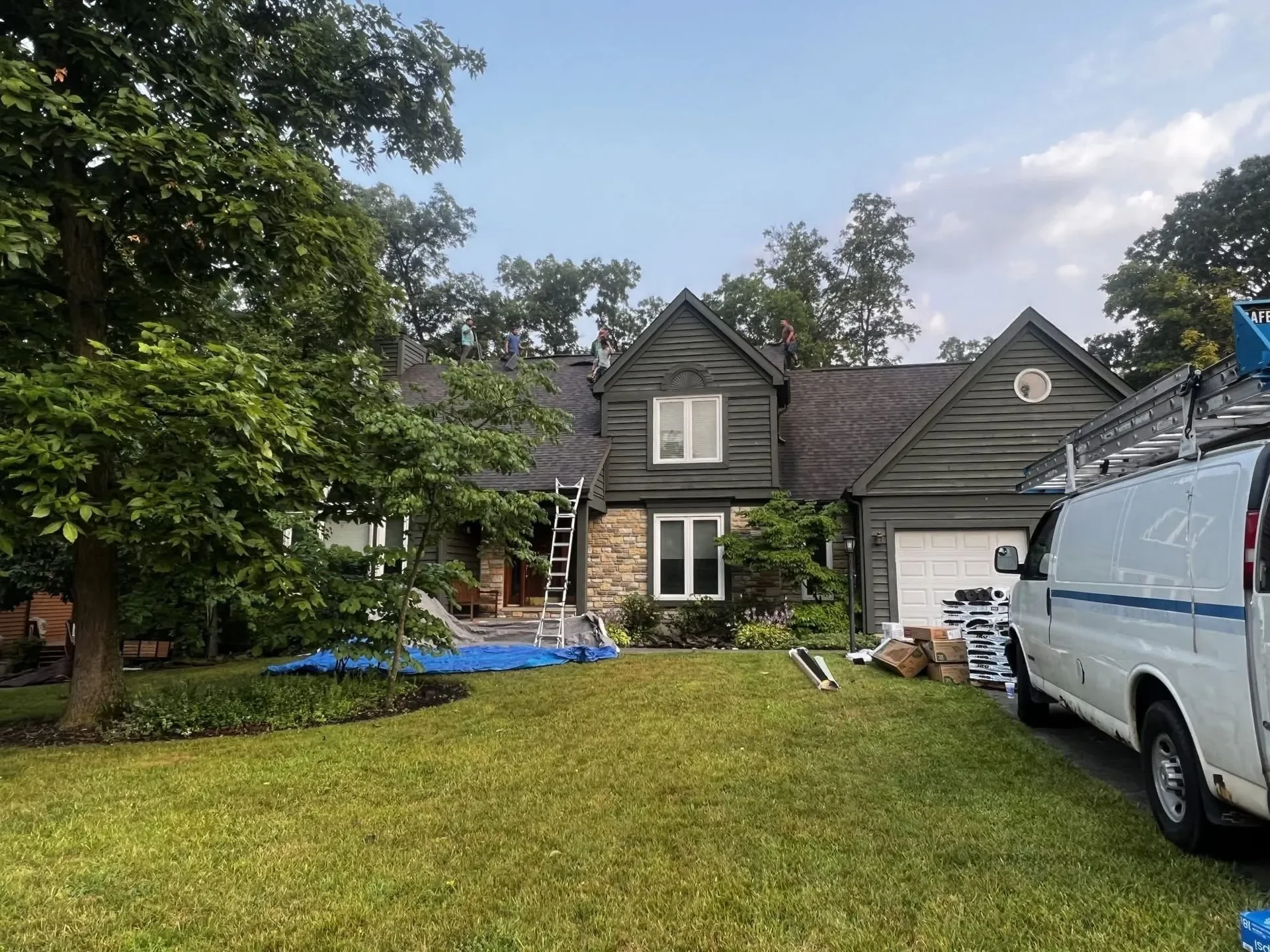A house with workers on the roof using safety harnesses, a ladder leaning against the house, a tree in the front yard, and a white van parked on the driveway with construction supplies around it.