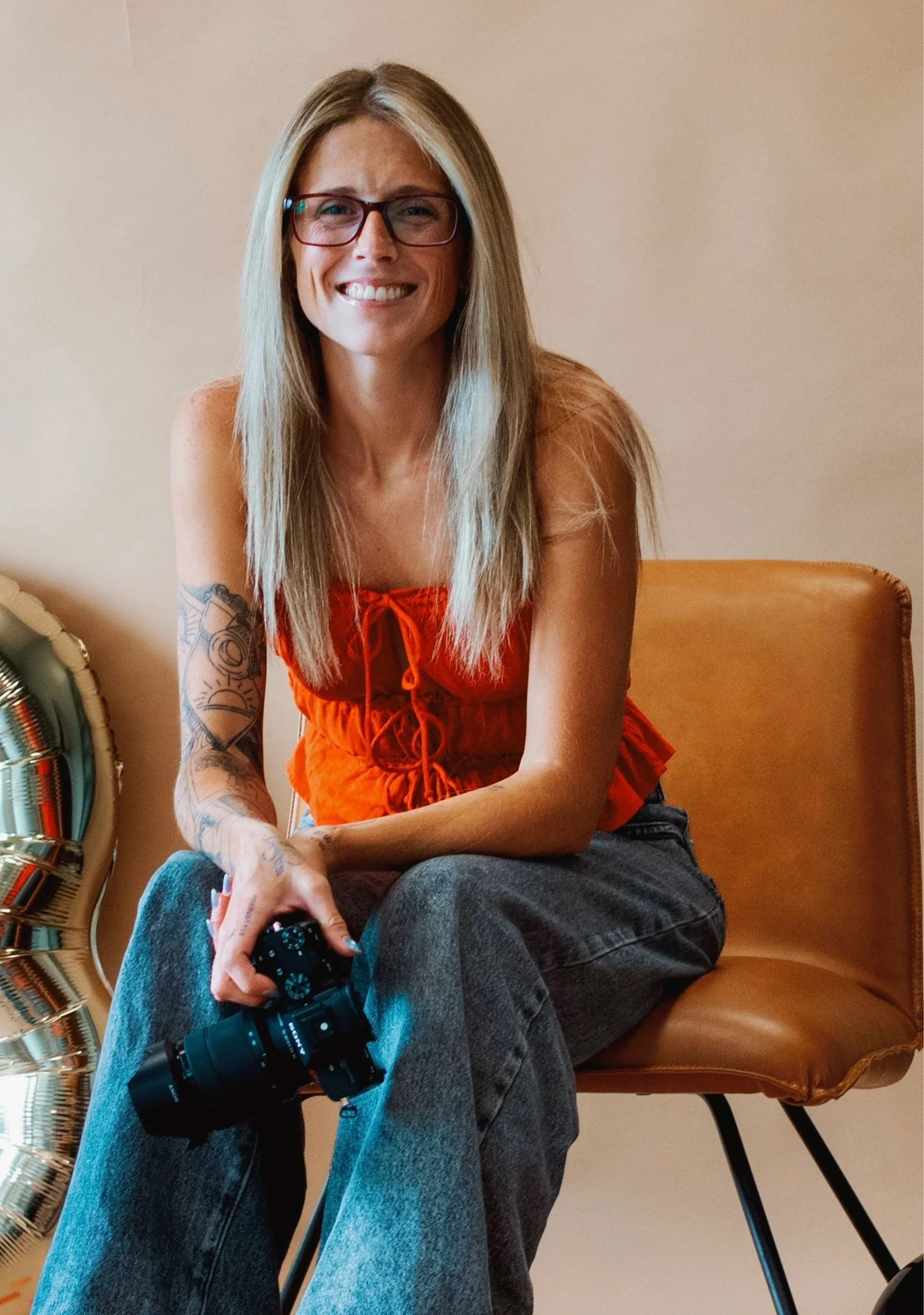 A woman with long blonde hair, glasses, and tattoos, smiling and holding a camera, sitting on a tan chair against a beige wall.
