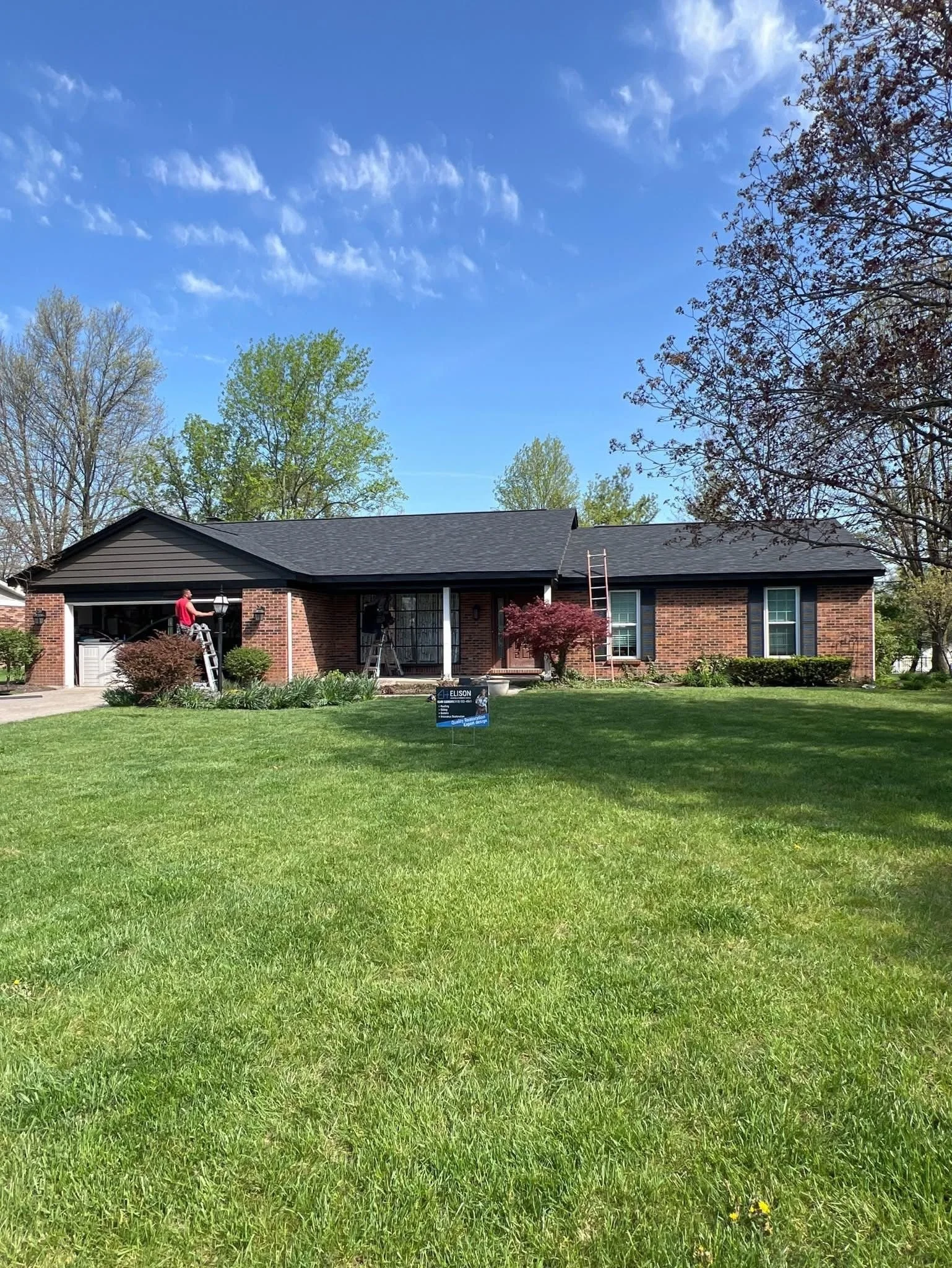 A brick house with a black roof and black shutters on the windows. There are two people working on ladders on either side of the house, possibly doing maintenance or repairs. The house has a front yard with green grass, small bushes, and trees. The s