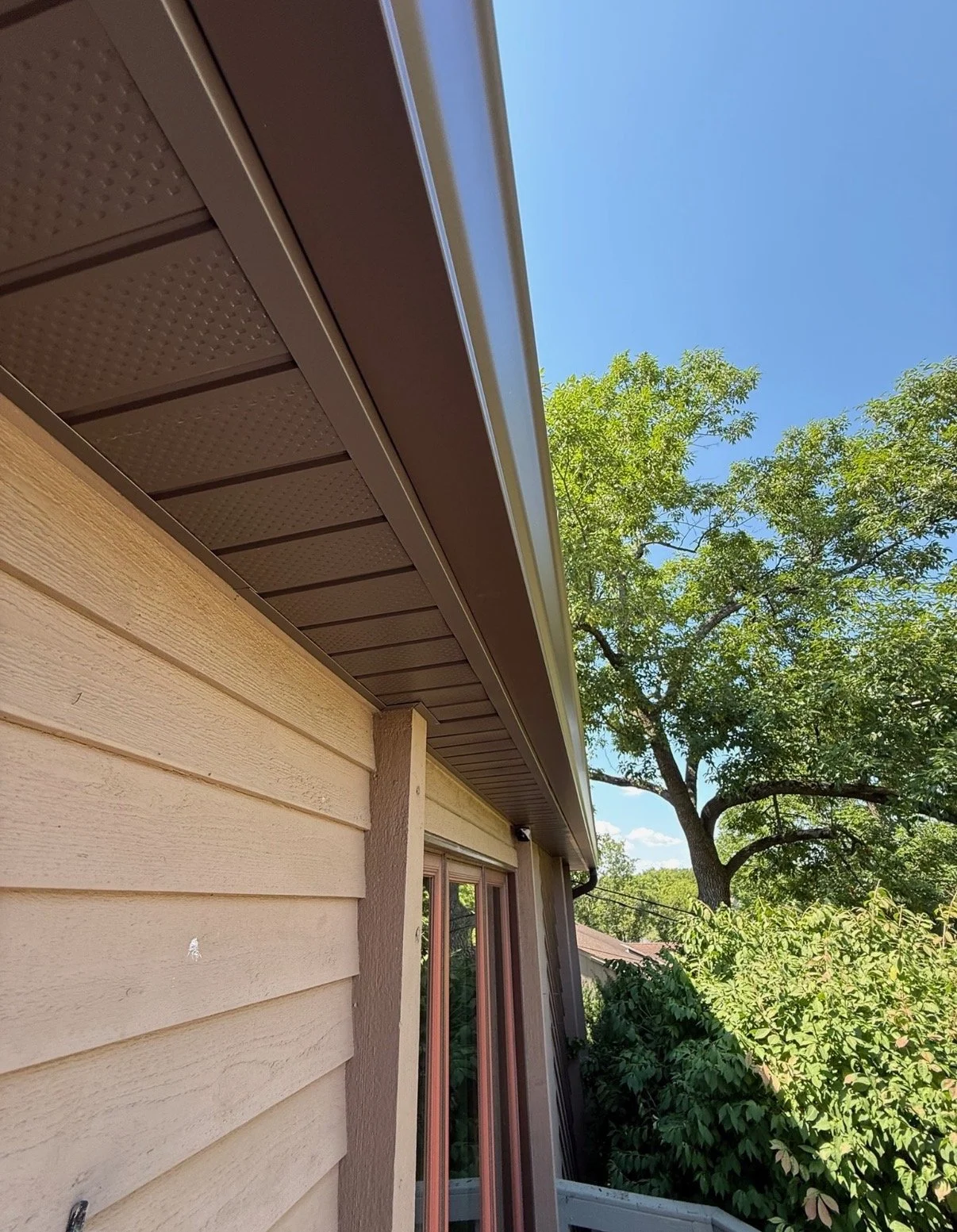 View of a house siding, eaves, and gutter with a tree and blue sky in the background.