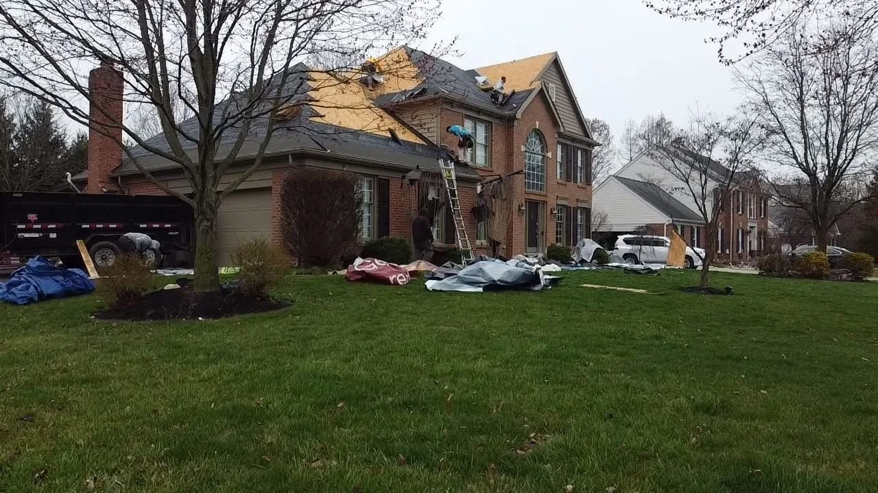 A house under construction with a new roof being installed. Workers are on the roof handling materials, and a ladder is leaning against the house. Trees and other houses are visible in the background.