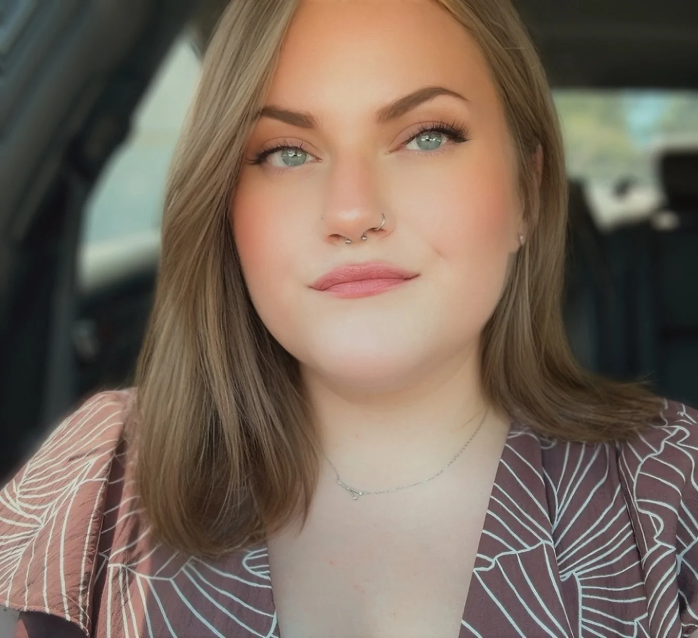 A young woman with shoulder-length brown hair, blue eyes, and a septum piercing, sitting inside a car with a blurred outdoor background.