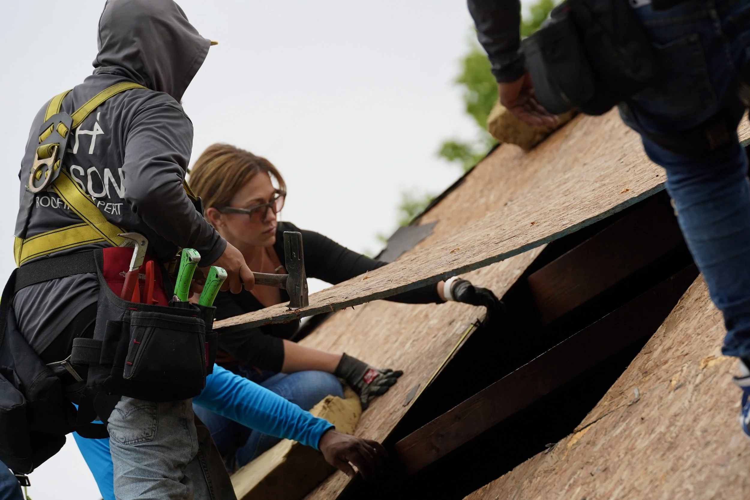 Two workers installing a roof, one hammering in nails and the other holding a wooden panel, with tools attached to their belts.