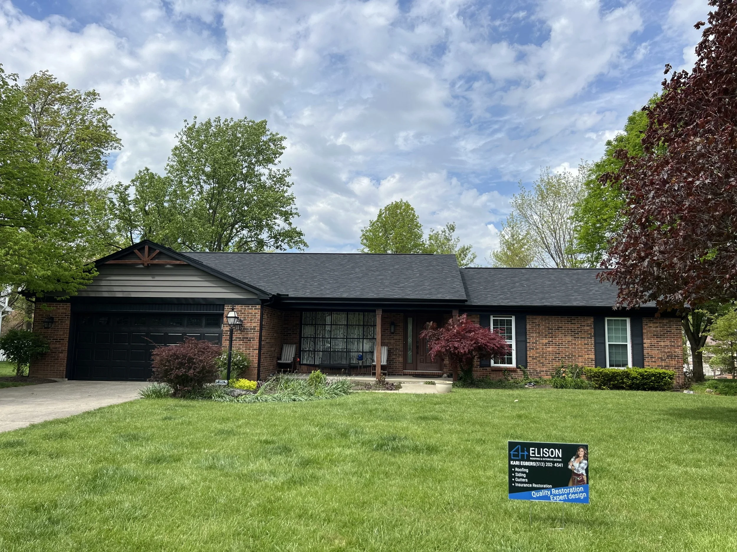 Single-story brick house with black garage door and black shutters, surrounded by green trees and a well-maintained lawn, with a sign in the yard advertising a roofing and restoration company.