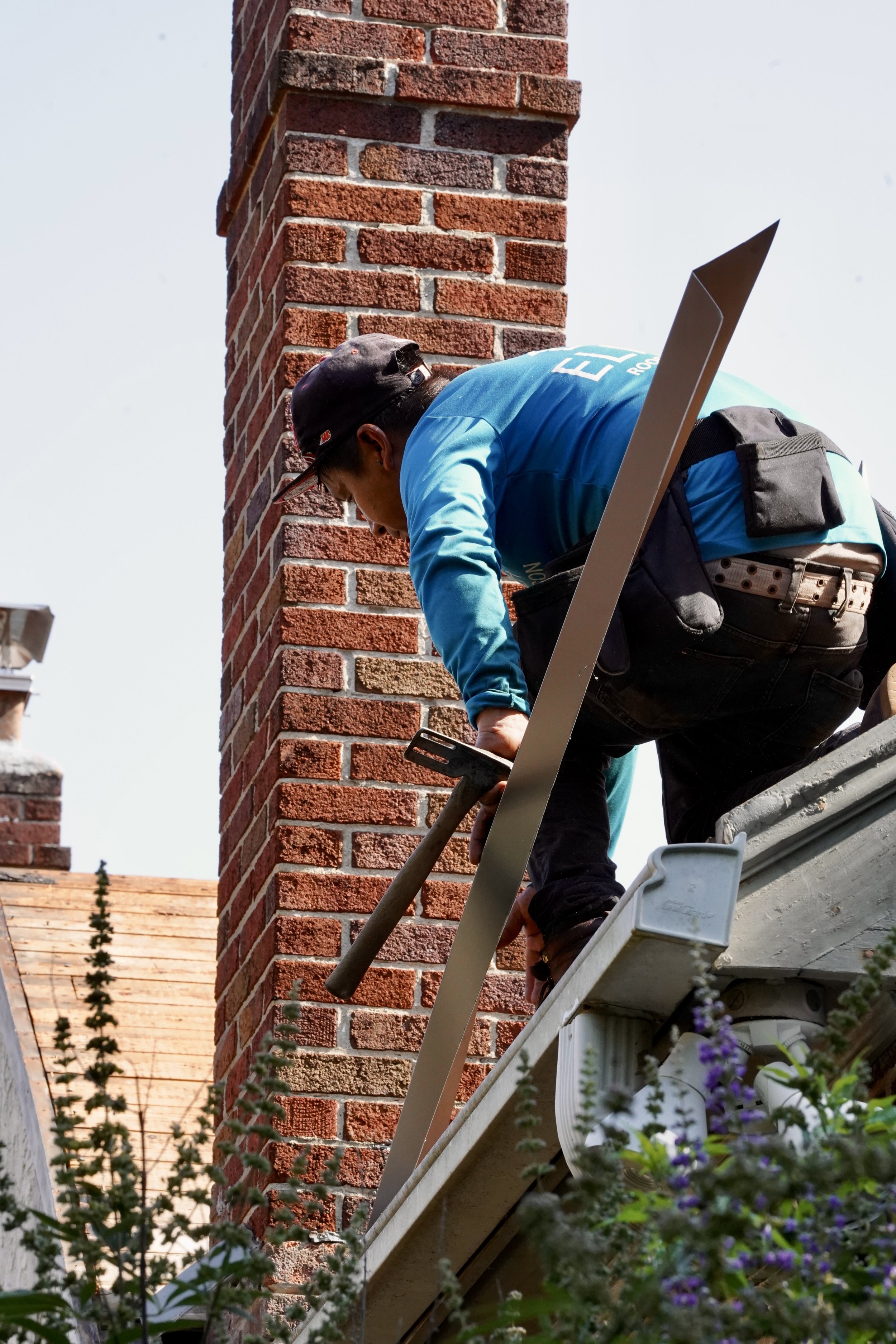 A worker installing or repairing a chimney on a house roof, holding a hammer, with some purple flowers in the foreground.