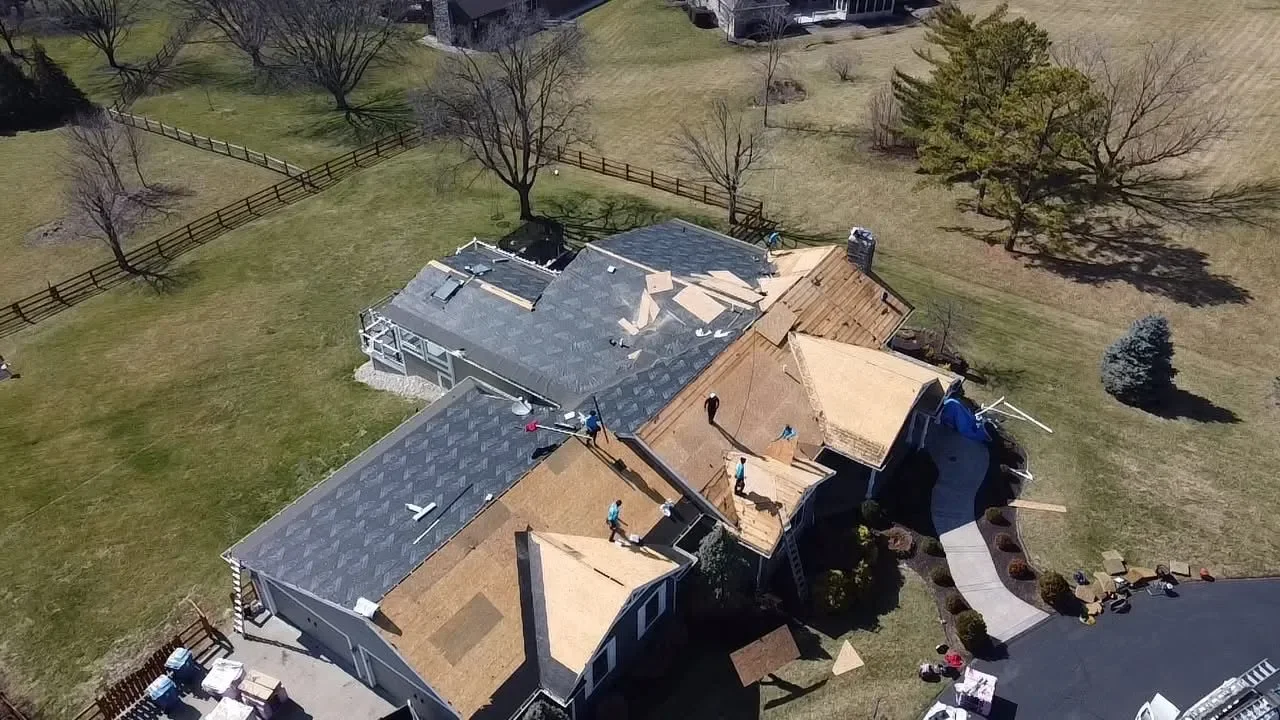 Aerial view of a house under construction with workers on the roof, with some sections of the roof being renovated or replaced, in a suburban backyard with trees and a fence.