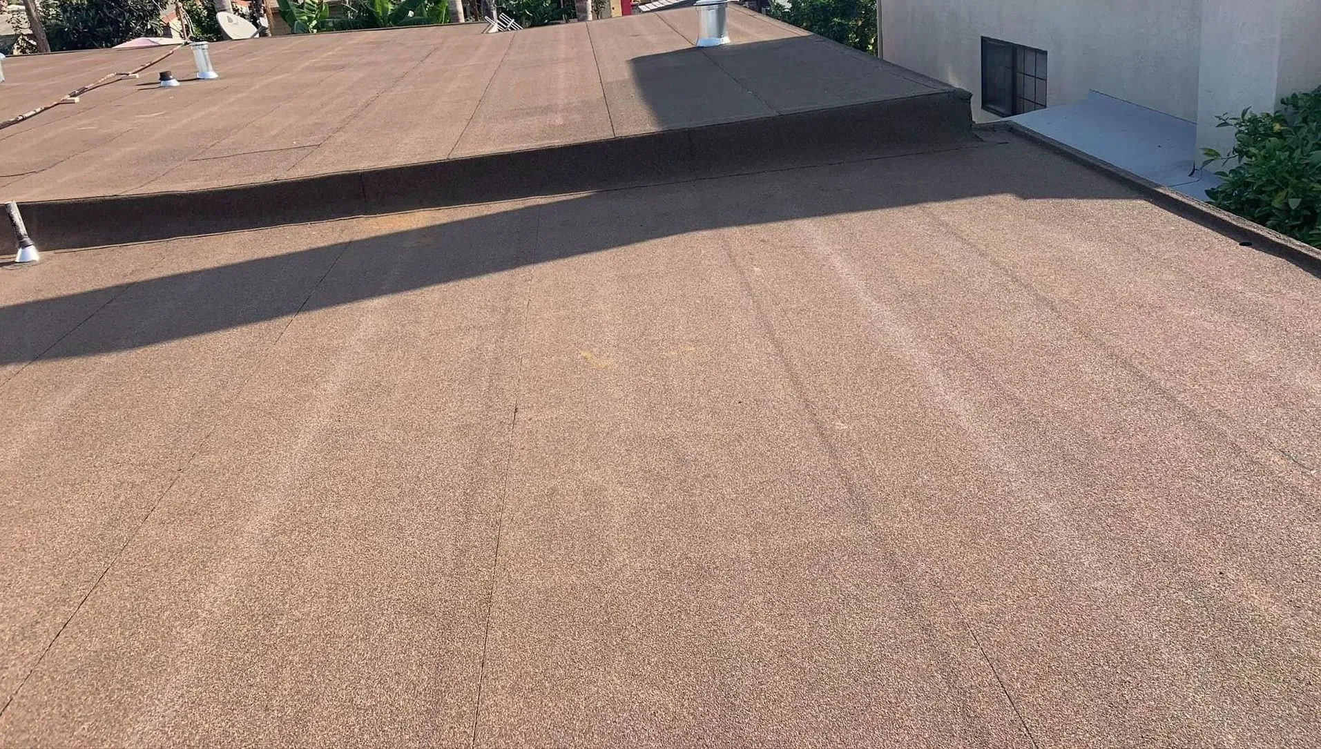 Flat rooftop with a brown surface, two vents, and a shadow cast across part of the roof. In the background, there is a building with a small window and some greenery.