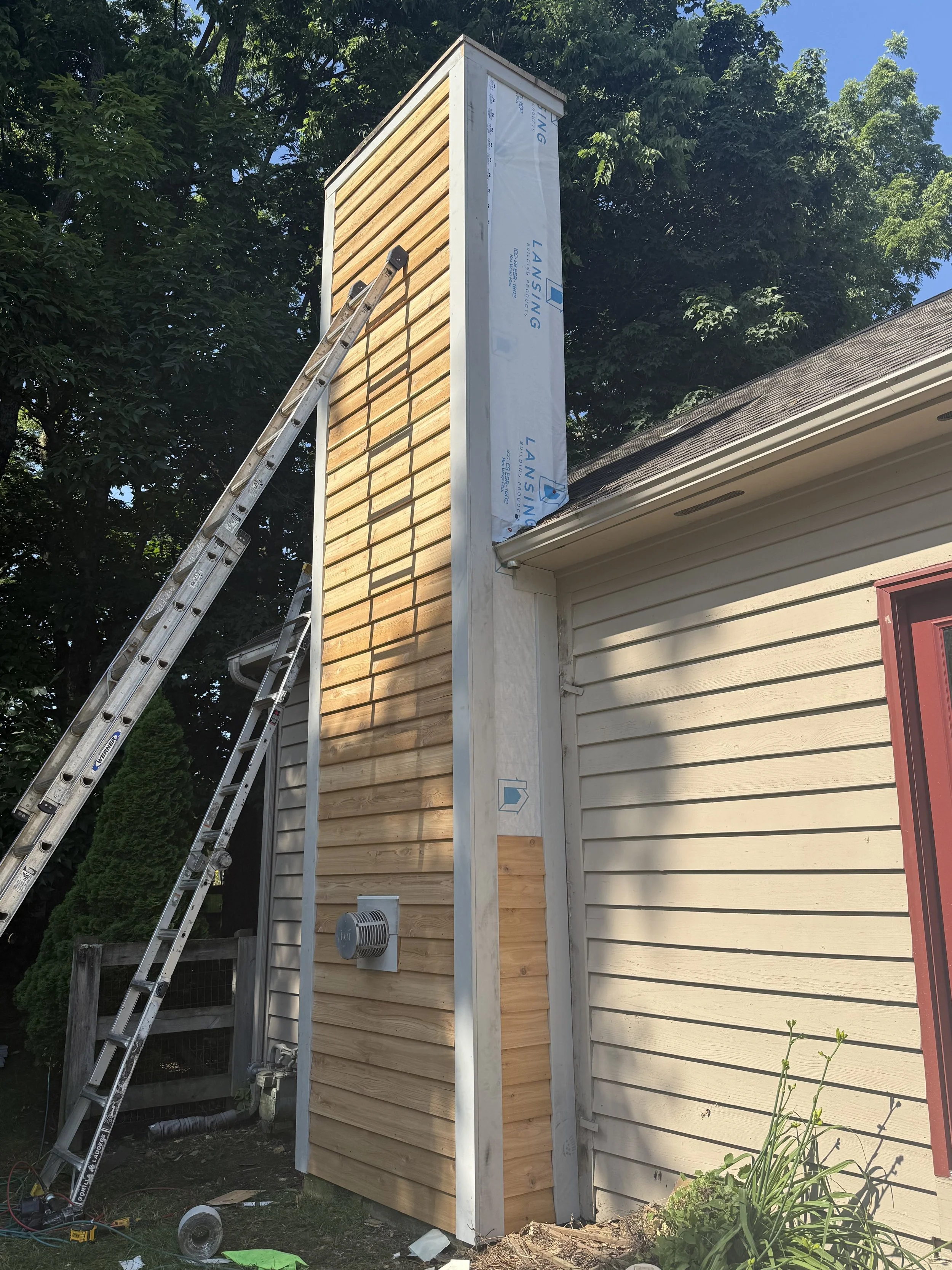 Construction work on a house chimney with wooden siding, ladders, and tools nearby.