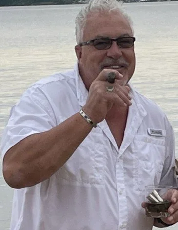 A man with white hair and glasses smiling, holding a cigar in his hand, wearing a white short-sleeve shirt with a flag patch, outdoors near water.
