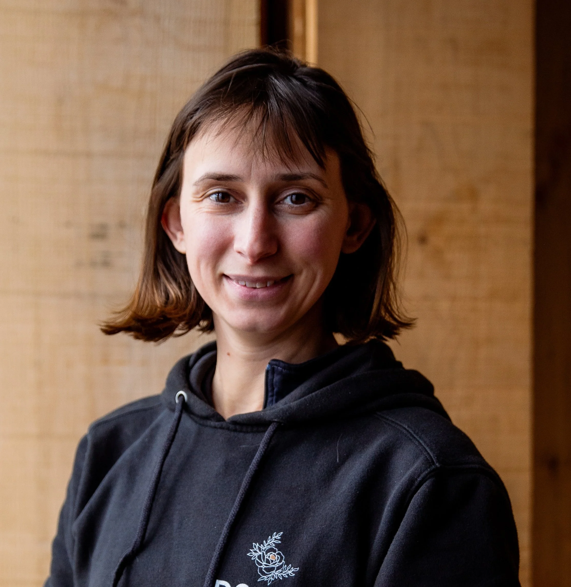 A woman with short brown hair wearing a black hoodie, smiling, indoors with wooden wall background.