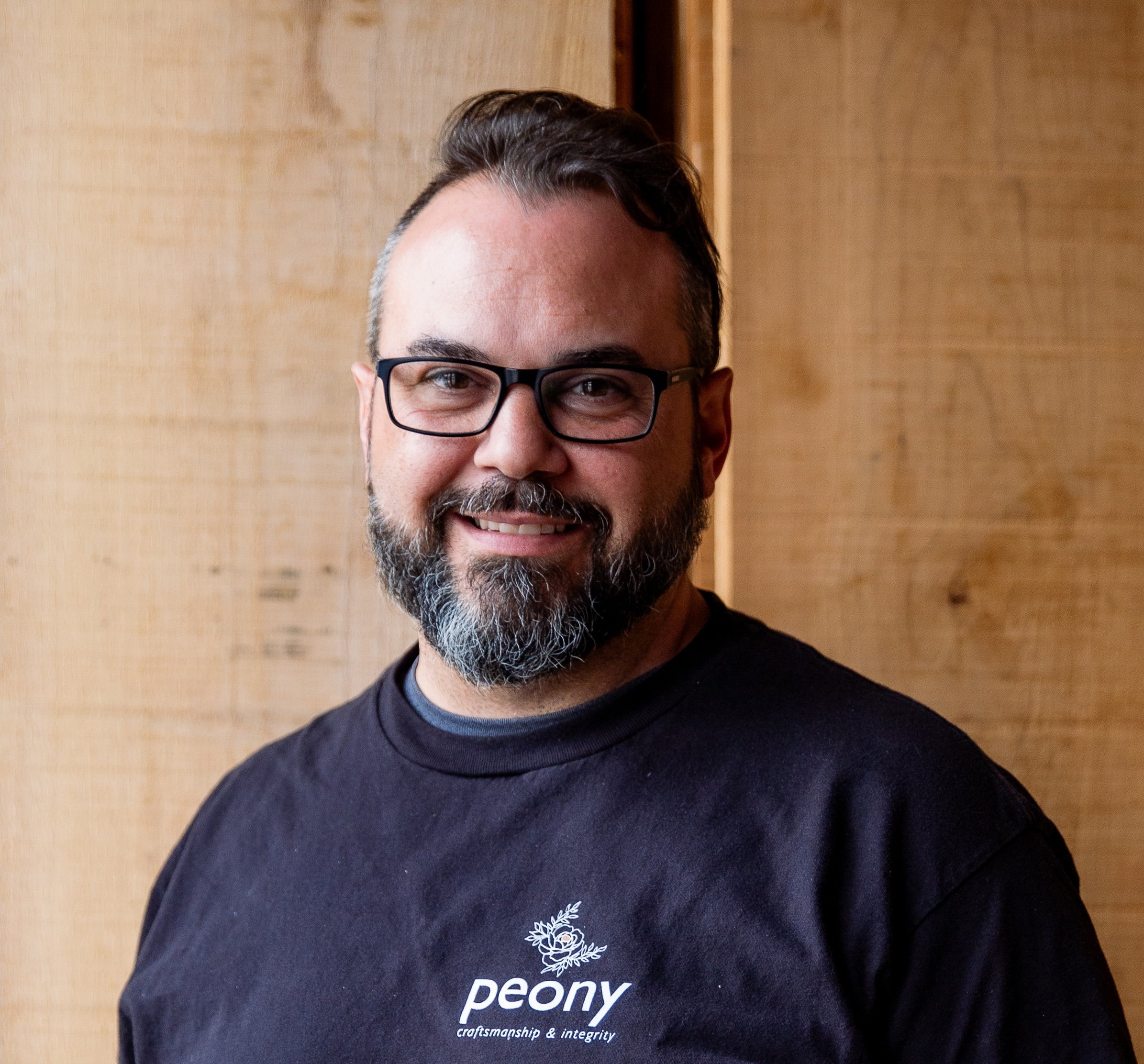 A smiling man with glasses, a beard, and short hair, wearing a black t-shirt with a peony design and the words 'craftsmanship & integrity.' standing in front of a wooden wall.