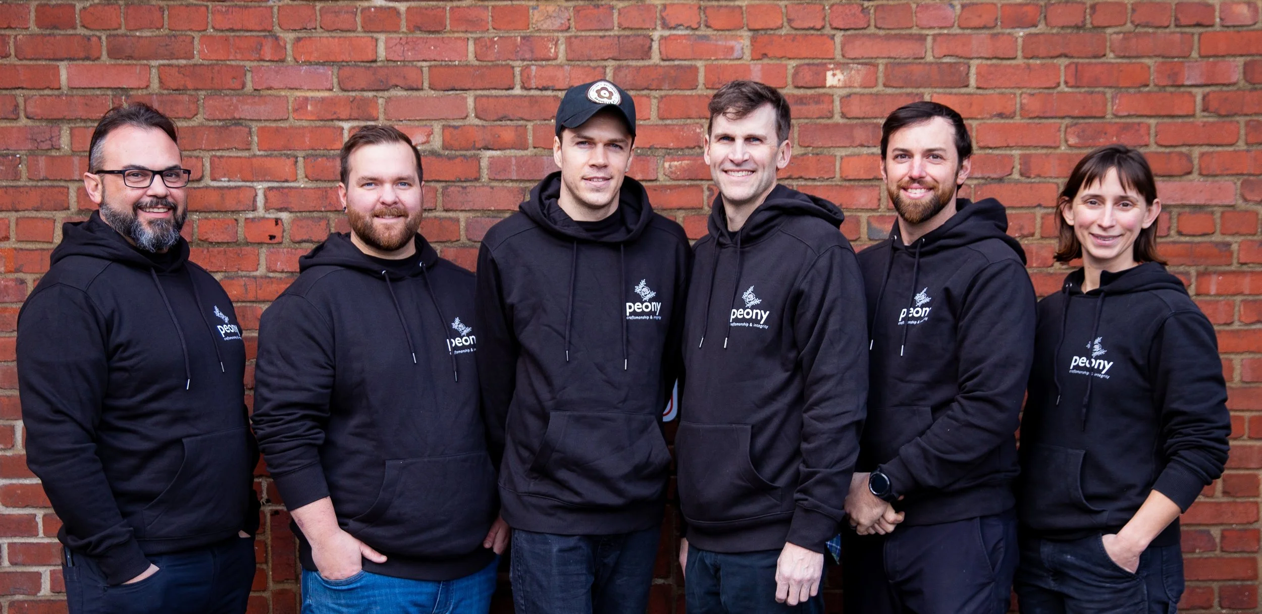 Group of six people standing in front of a brick wall, wearing black hoodies with a logo that reads "peony".