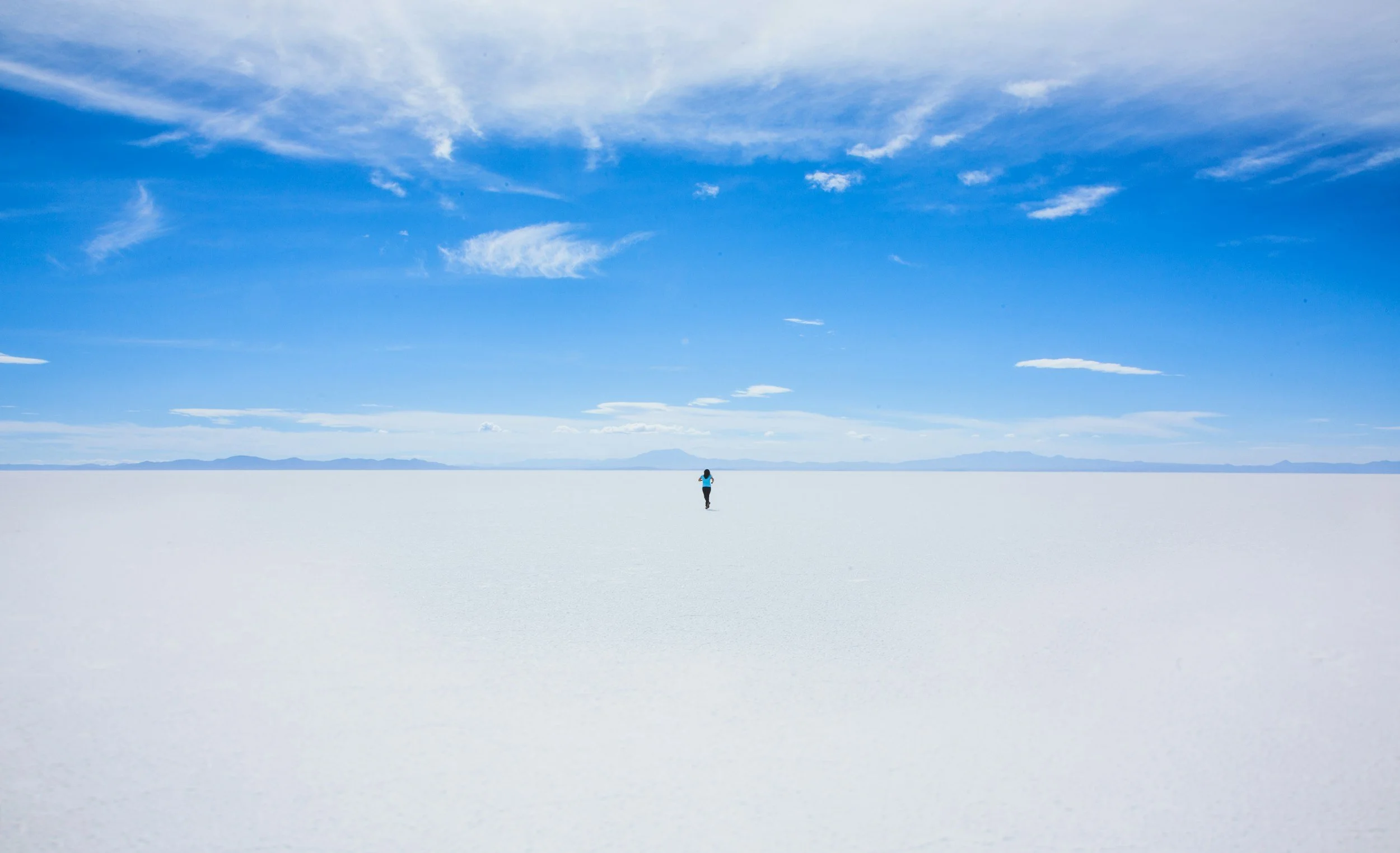 A person standing alone on a vast, flat white salt flat under a blue sky with scattered clouds and distant mountains.