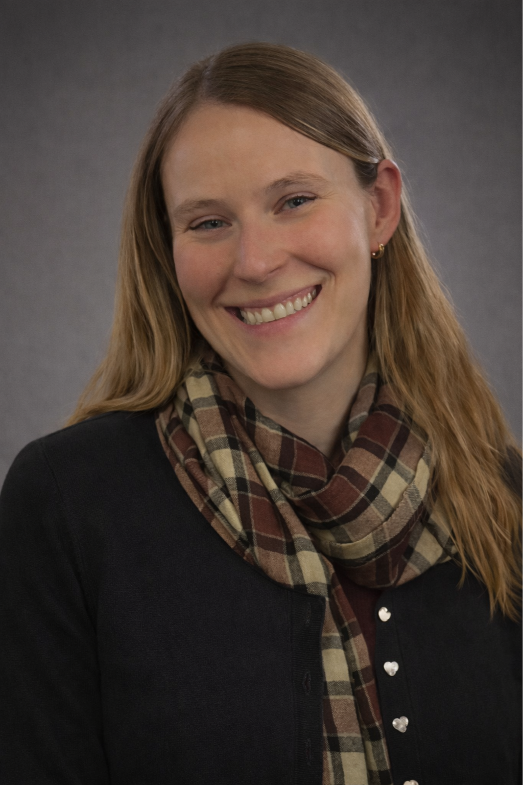 A woman with red hair and blue eyes smiling, wearing a black sweater and a beige, brown, and red plaid scarf, against a dark grey background.