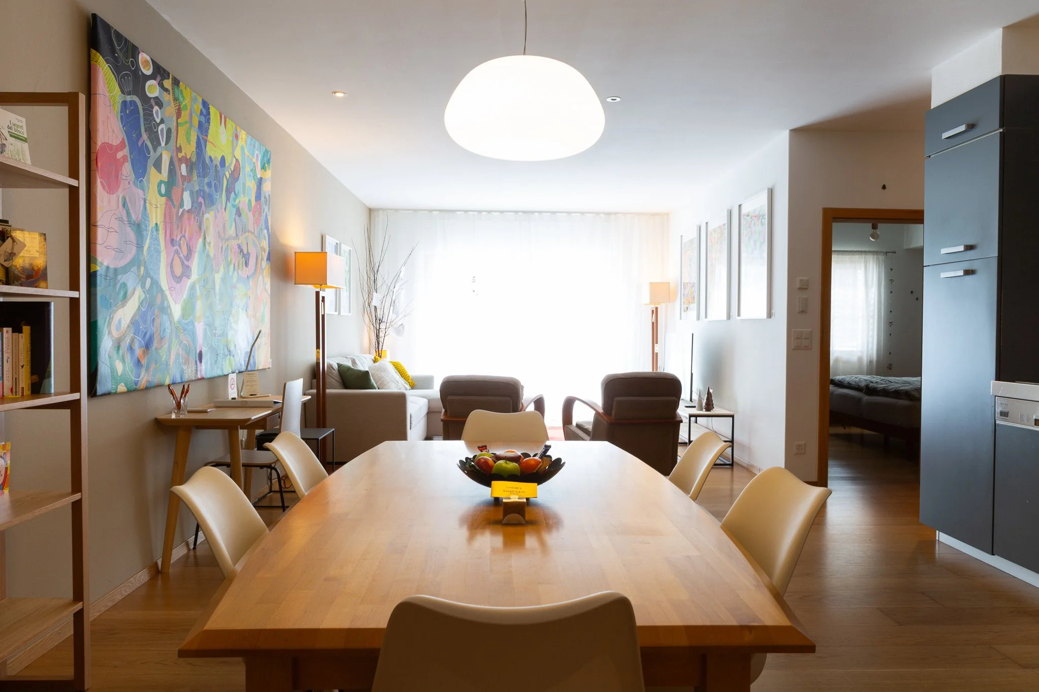 Sala da pranzo con tavolo di legno e sedie chiare, soggiorno con divani grigi, tavolino e lampade, pareti decorate con quadri, finestra grande con tende bianche, stanza da letto visibile attraverso la porta aperta, arredamento moderno e luminoso.