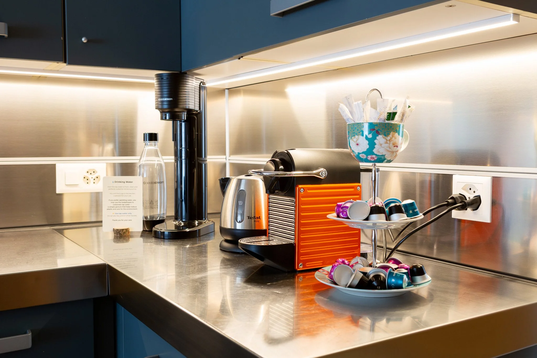 Angolo cucina con macchina da caffè, dispenser di acqua, cubetti di zucchero, tazza con bustine di tè o caffè, e recipiente con capsule di caffè.