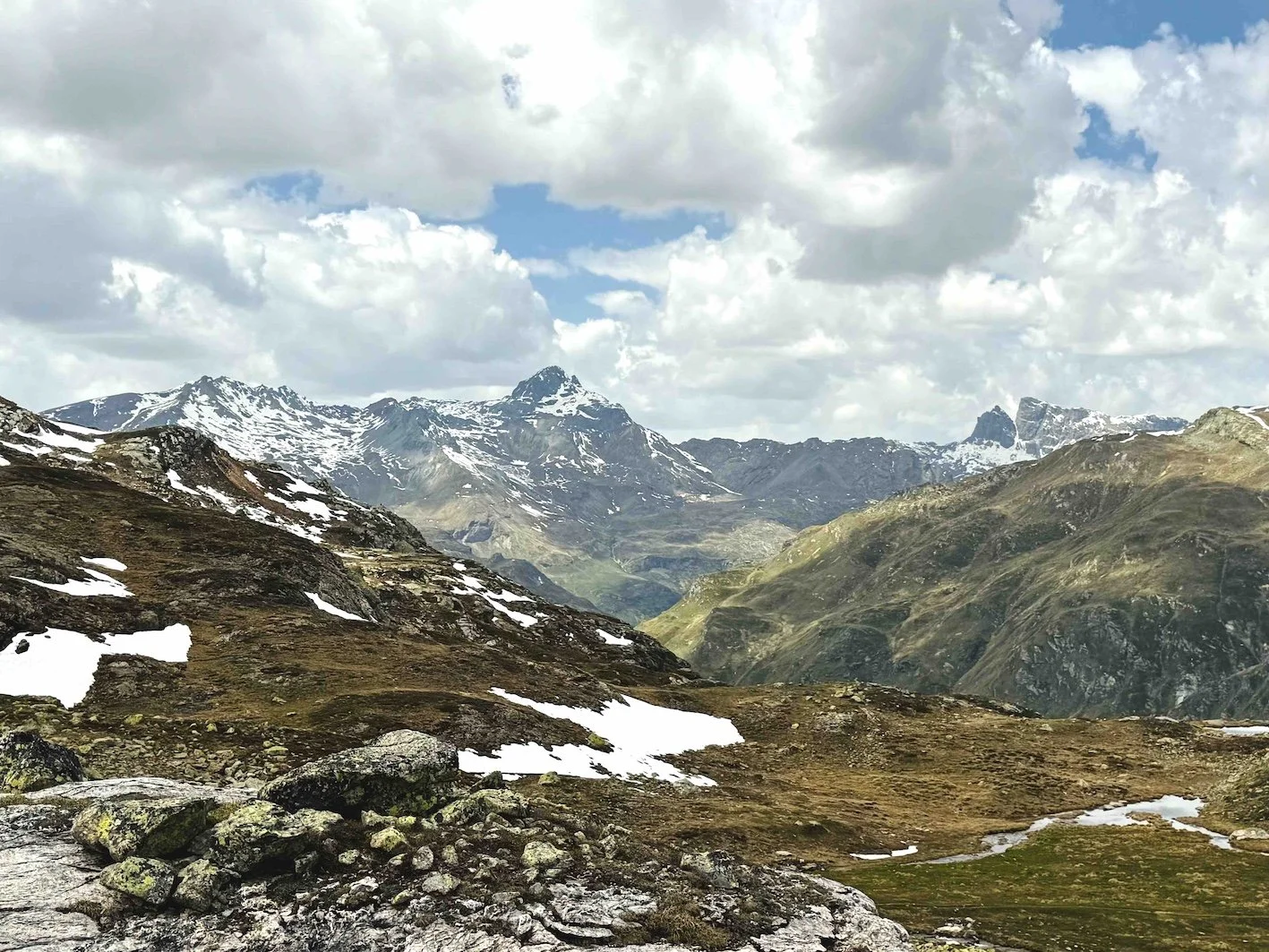 Paesaggio montano con cime innevate, nuvole e terreni rocciosi e verdi.