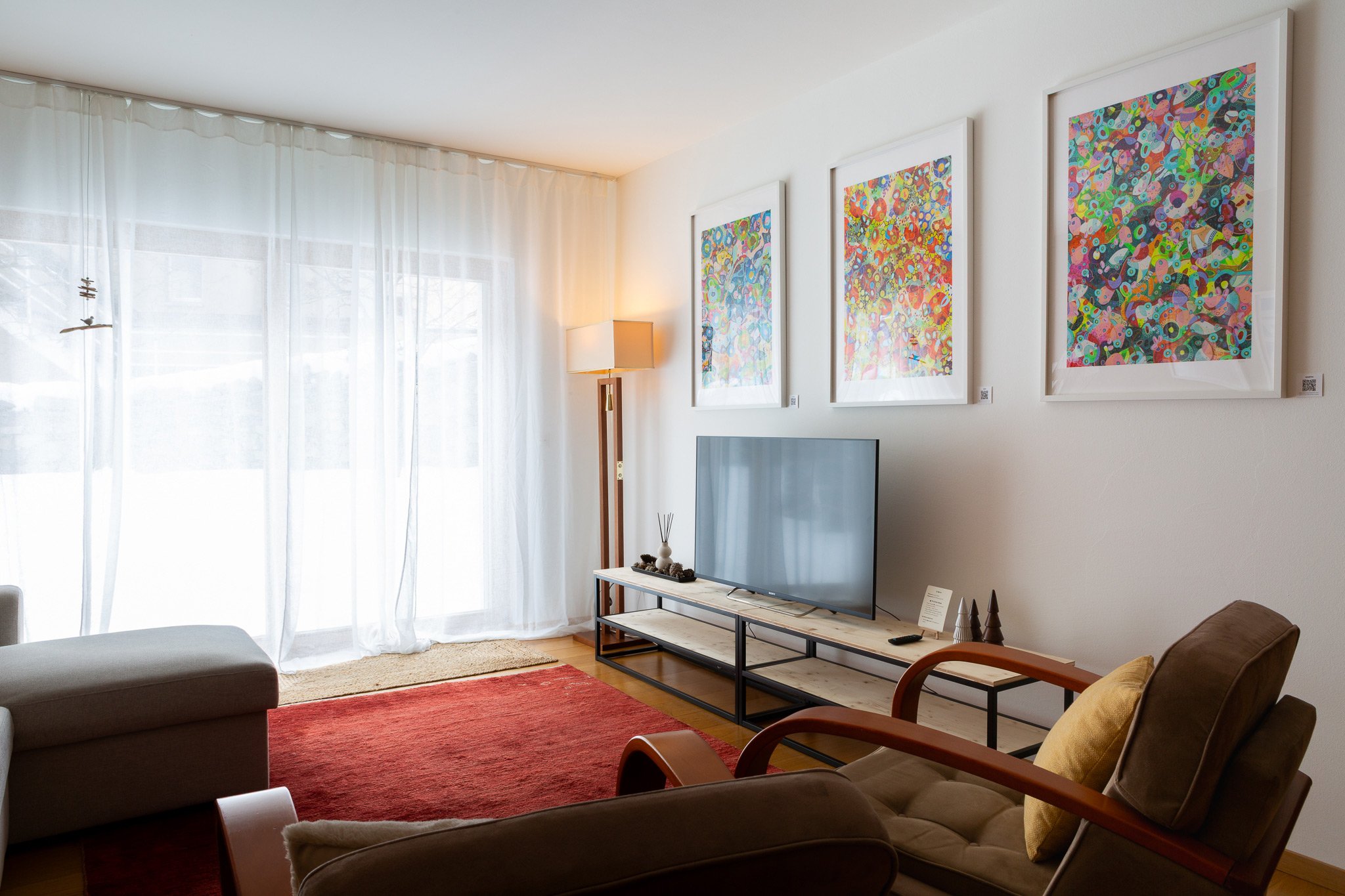 Soggiorno con parete bianca, tre quadri astratti colorati, grande finestra con tende bianche, tavolino TV con decorazioni e divani in stile moderno.