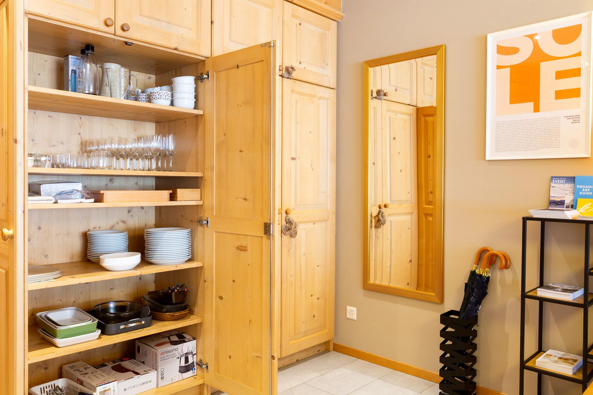 Armadio con piatti, bicchieri e ciotole, specchio e poster sulla parete, con ombrelli e riviste sulle mensole.