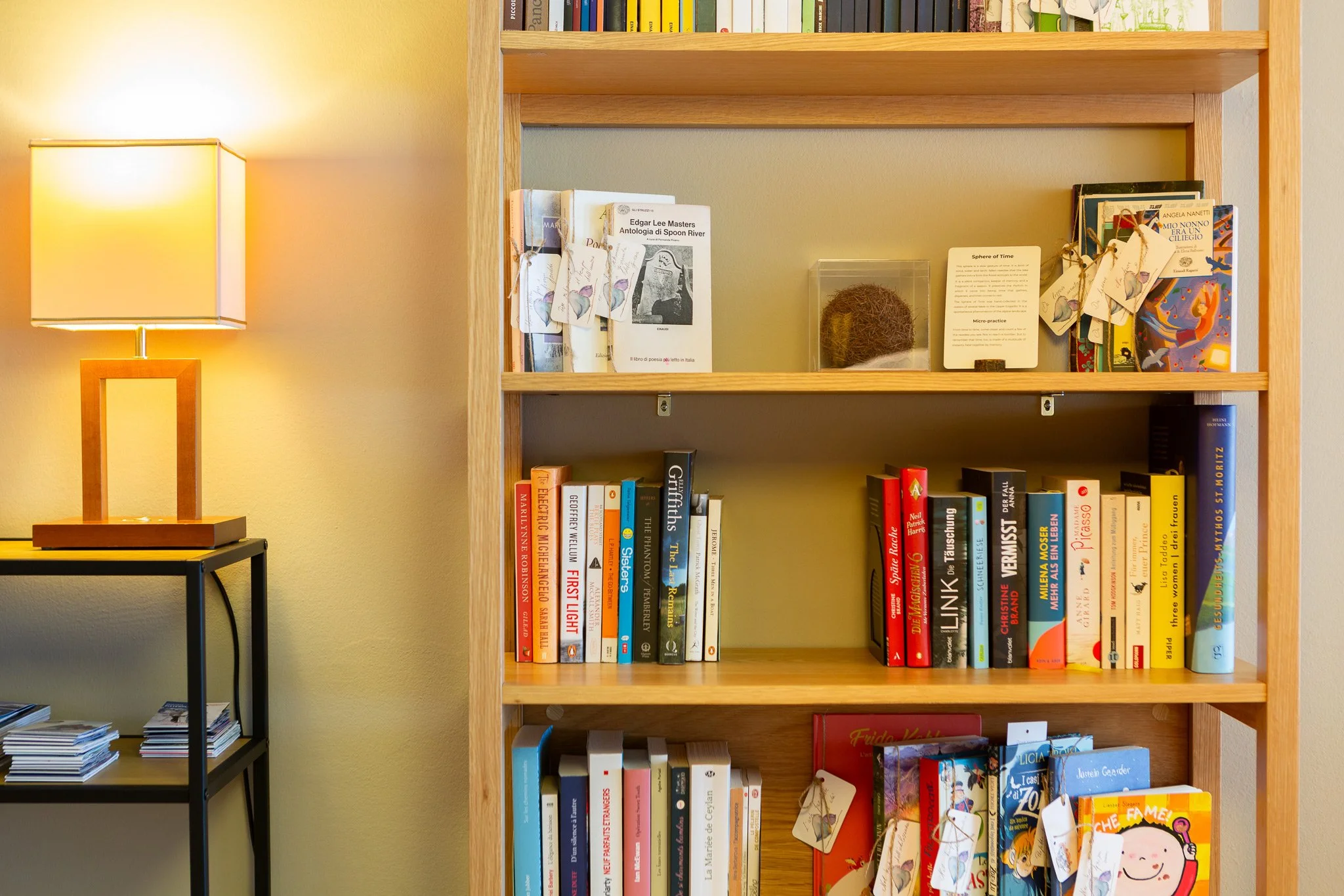 Scaffale di legno con libri di vari colori e dimensioni, alcuni con etichette di carta attaccate. A sinistra, lampada da tavolo su un tavolino nero, che emette luce calda.