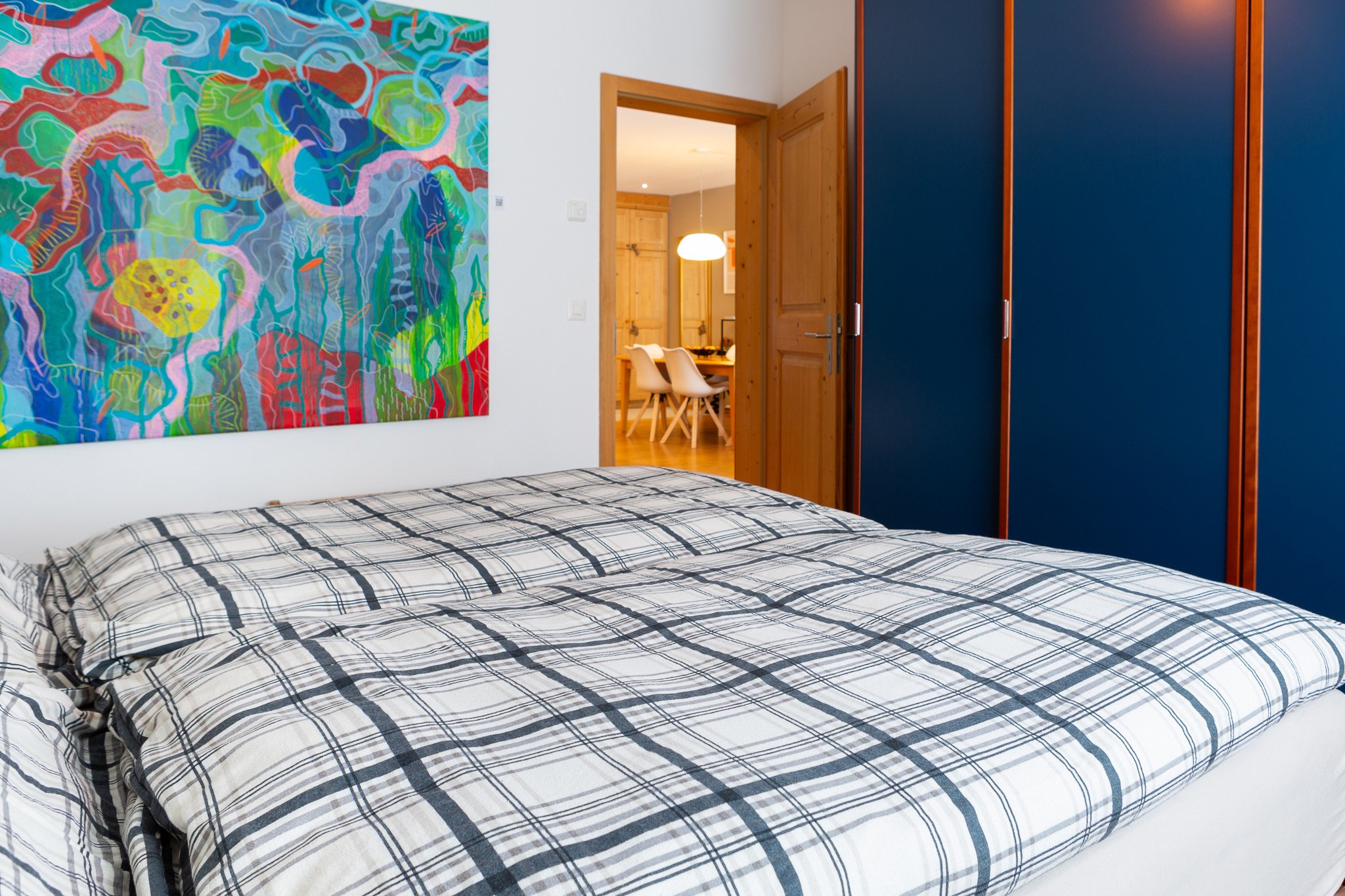 Camera da letto con letto in legno e copripiumoni a quadri, pareti bianche, arte astratta colorata sulla parete e armadio blu con dettagli in legno, vista di una sala da pranzo attraverso la porta aperta.