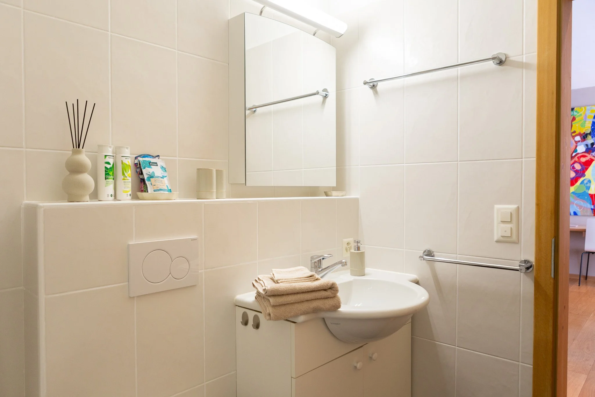 Bagno con lavabo bianco, specchio, asciugamani color beige, e vari prodotti per l'igiene sul ripiano.