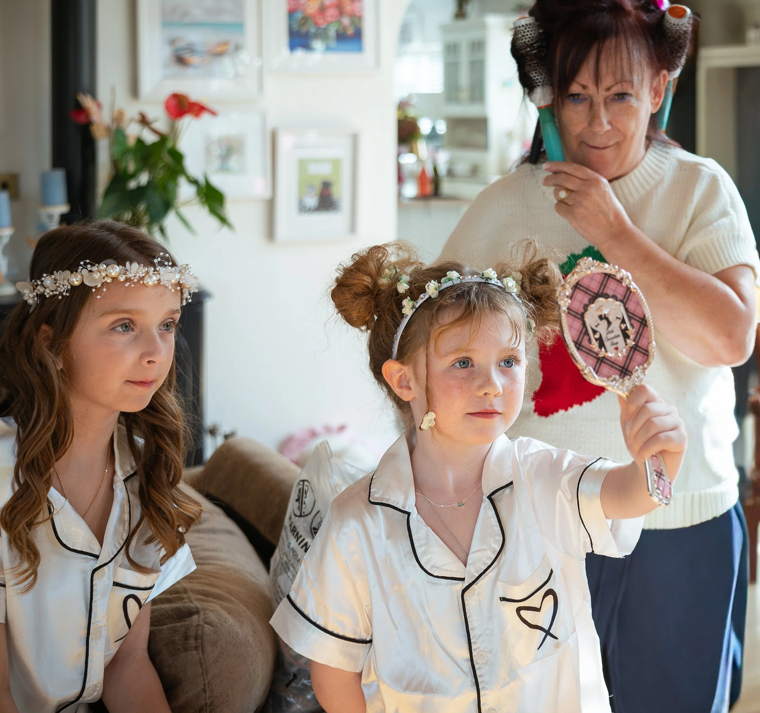 Three young girls with floral headbands in pajamas, standing in a room, one holding a mirror, while an older woman with curlers in her hair looks on.