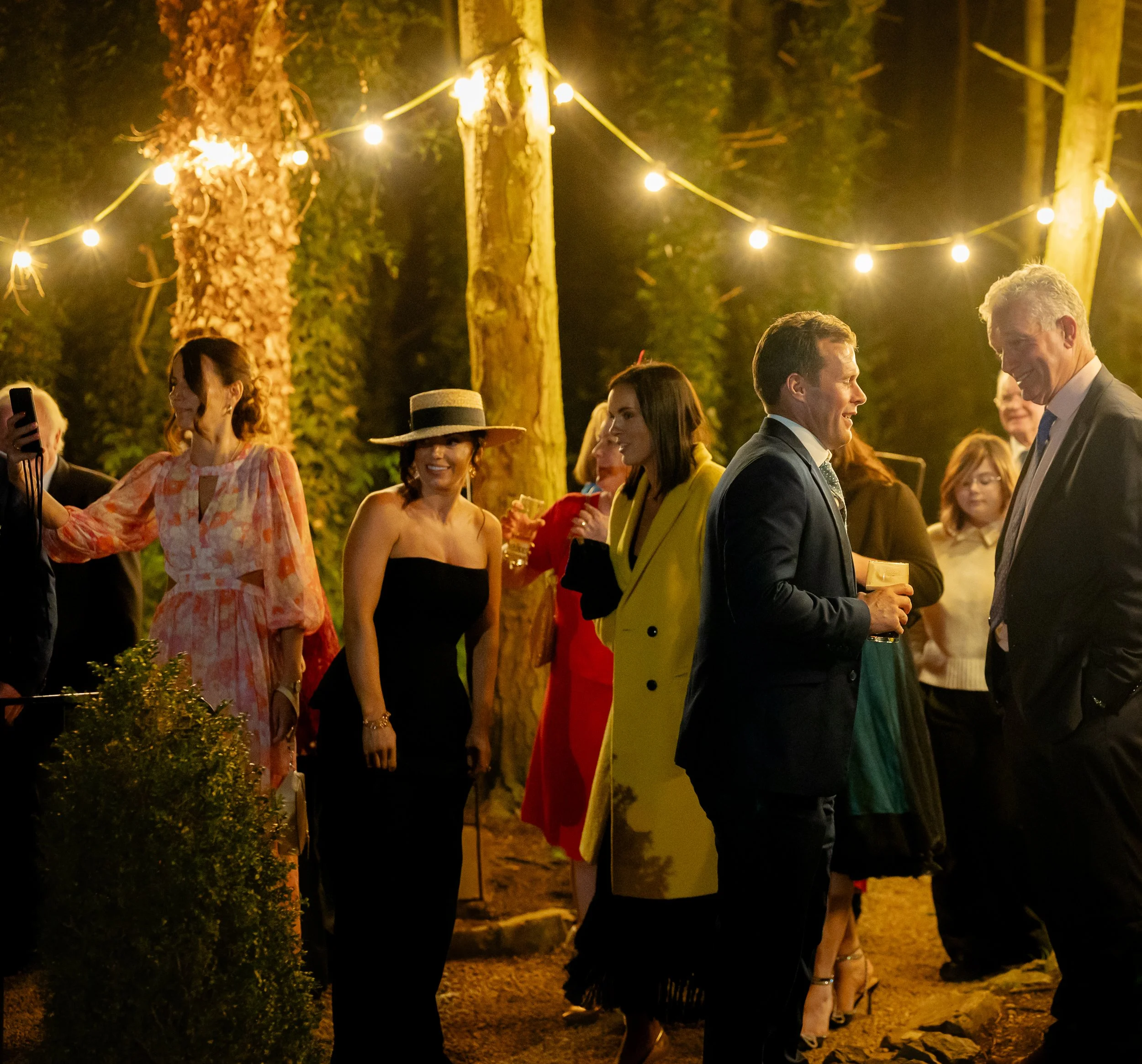 People enjoying an outdoor evening party, wearing formal attire, with string lights overhead in a wooded area.