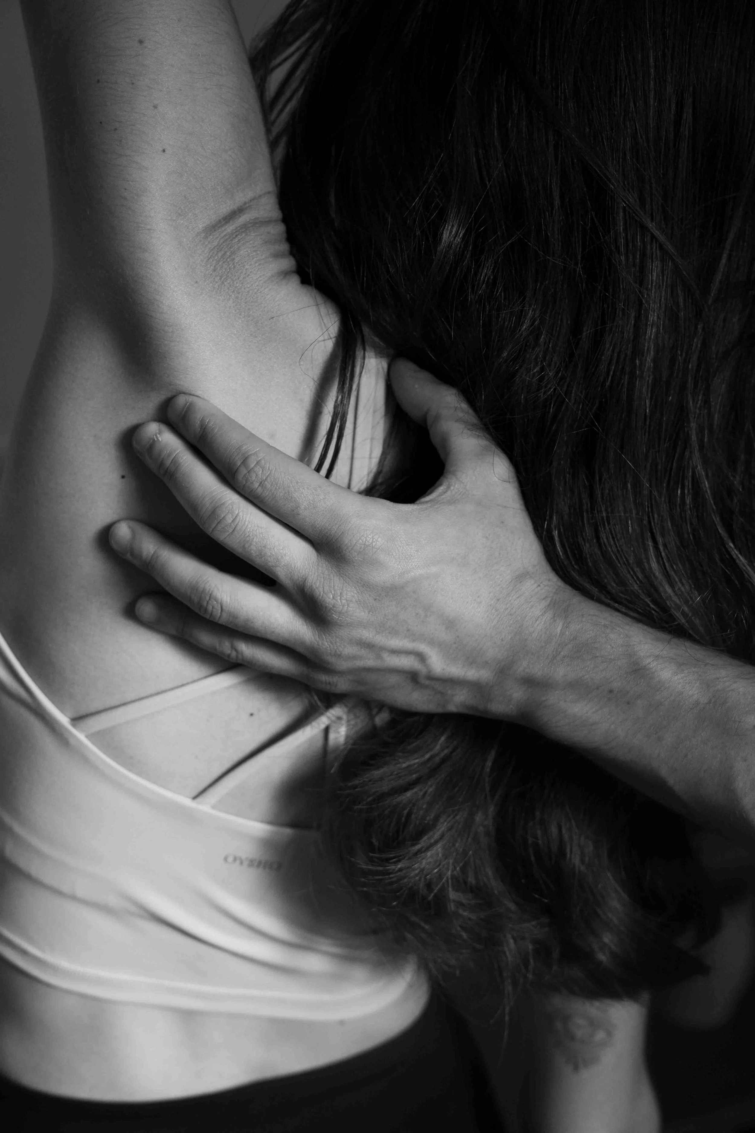 A person with dark hair covering their face, resting their head on their arm, which is raised and bent at the elbow. The image is in black and white.