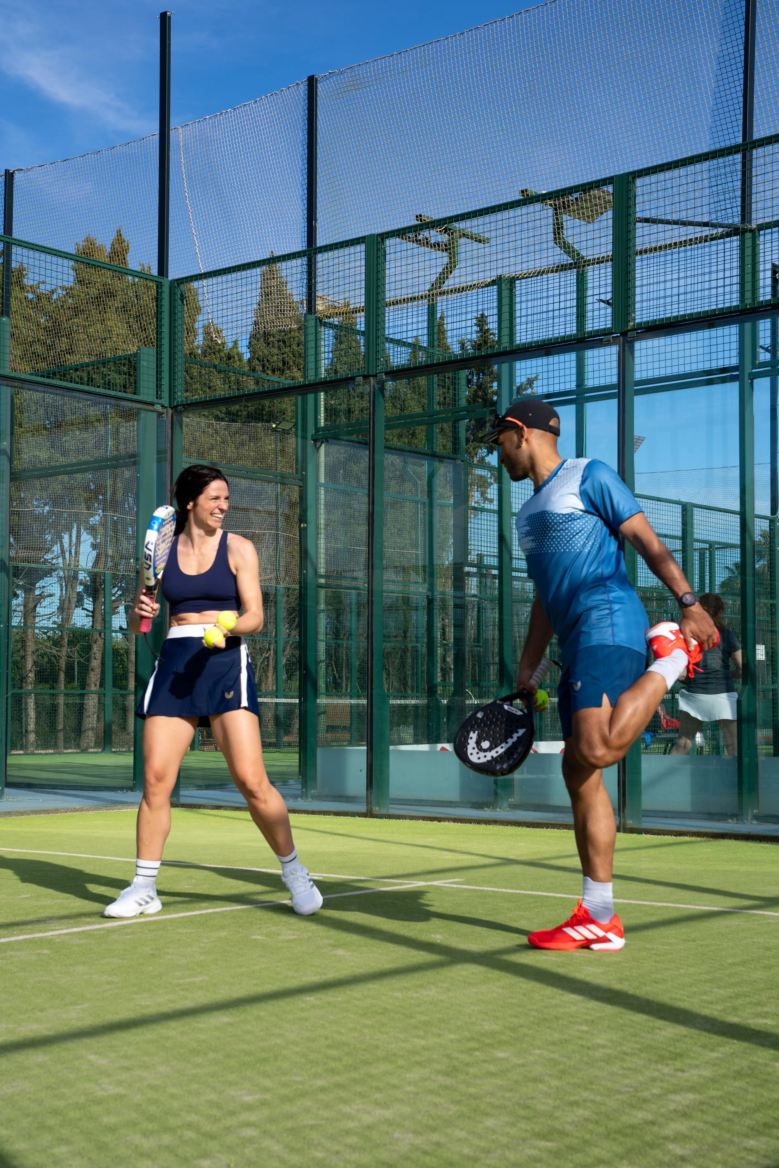 Two people playing paddle tennis on an outdoor court, holding paddles and tennis balls, with a blue sky and trees in the background.